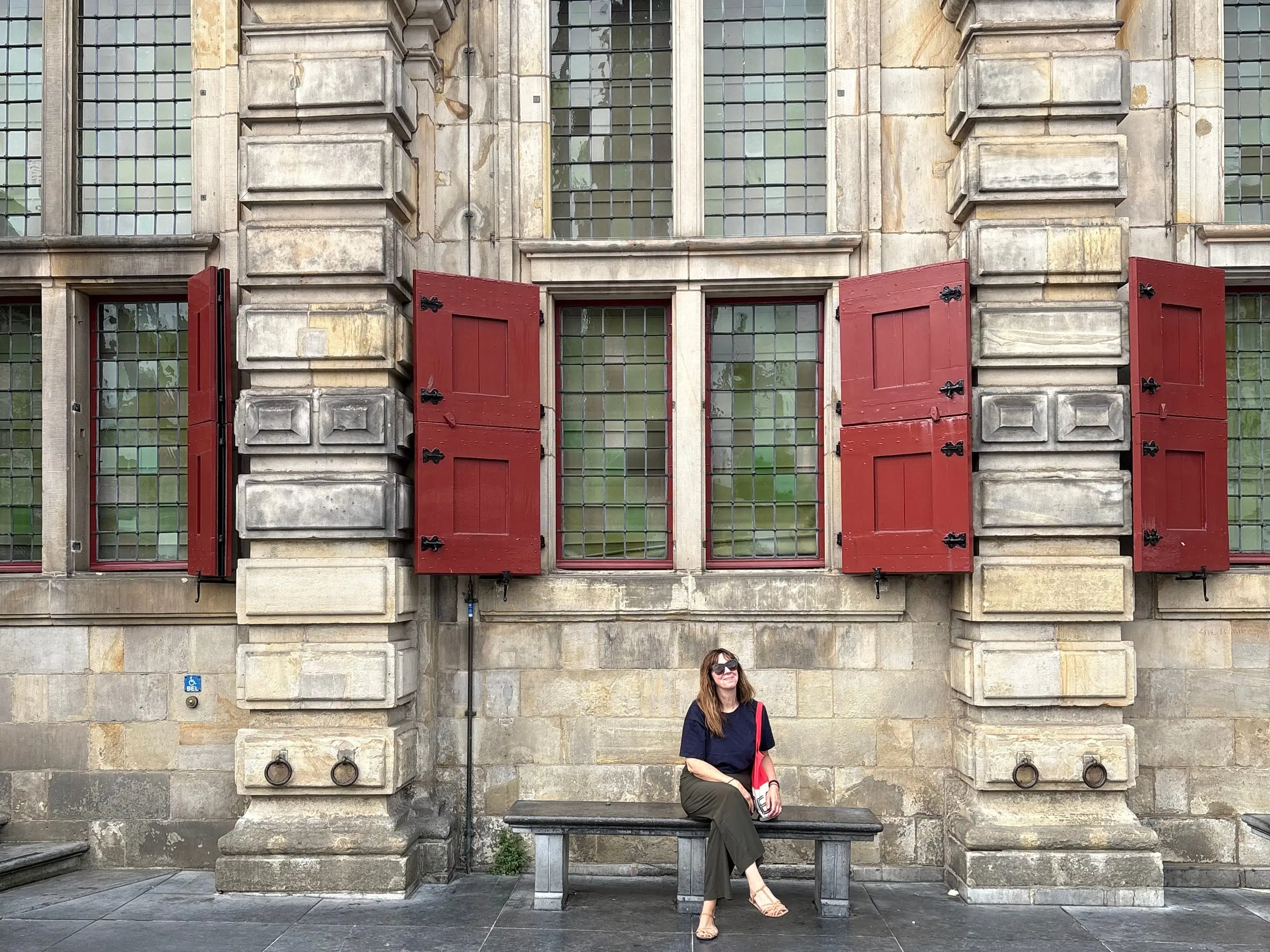 A seated person rests on a bench beneath red shutters on an ornate historic stone building facade.