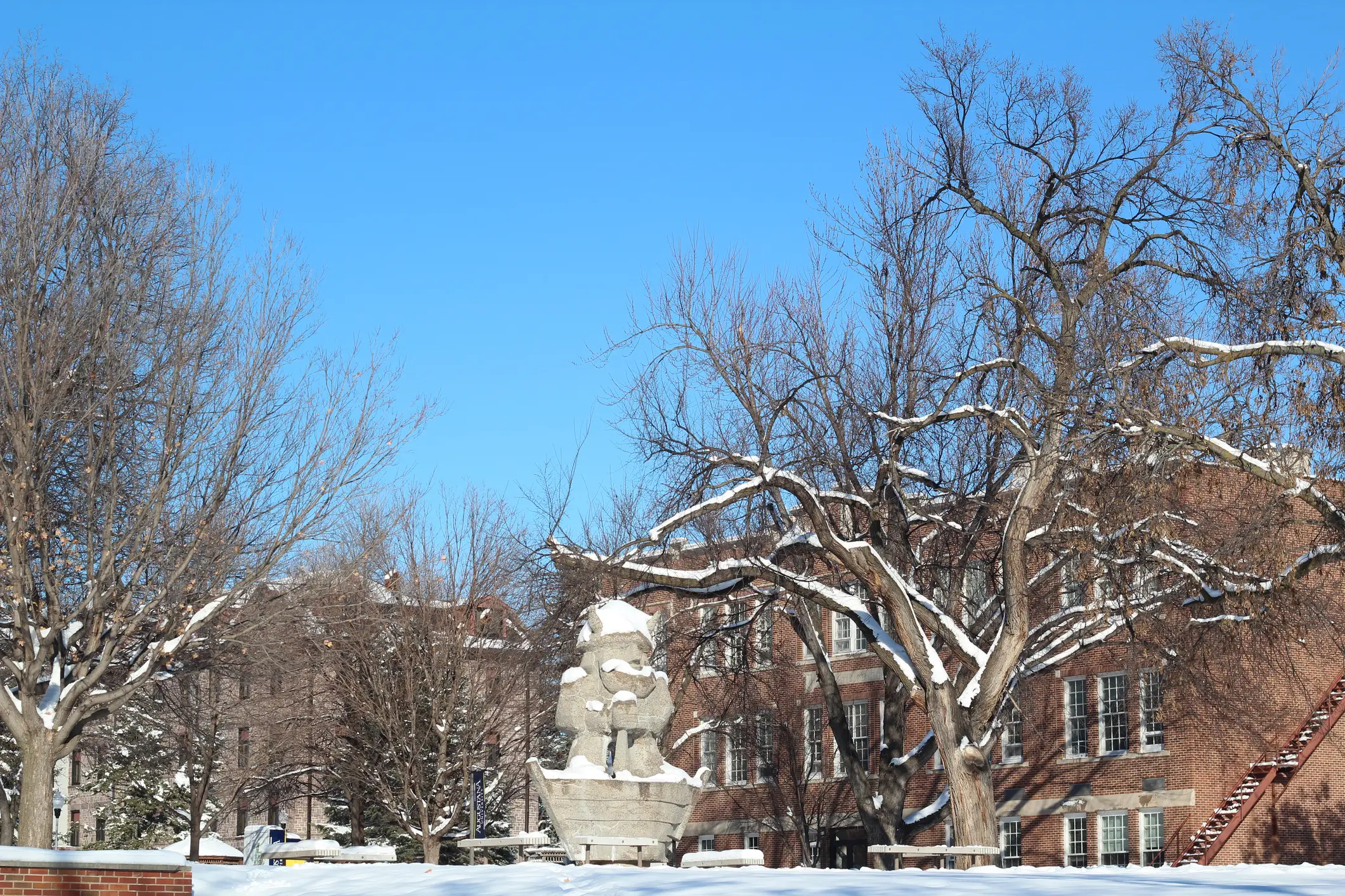 Snow-covered trees surround a stone fountain in front of brick campus-style buildings under a clear blue sky.