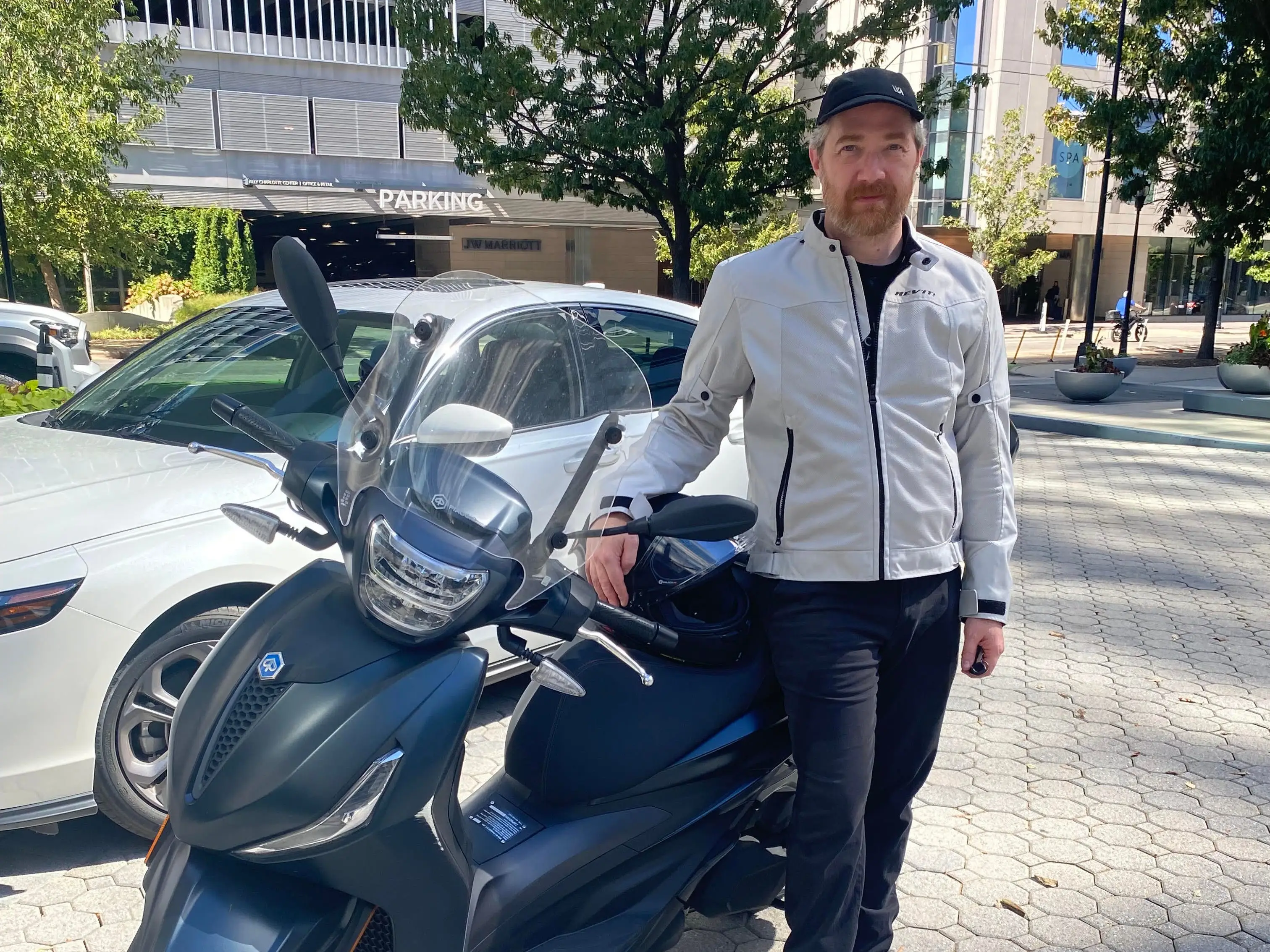 Person in a light jacket stands beside a dark Piaggio scooter on a city sidewalk near parked cars.