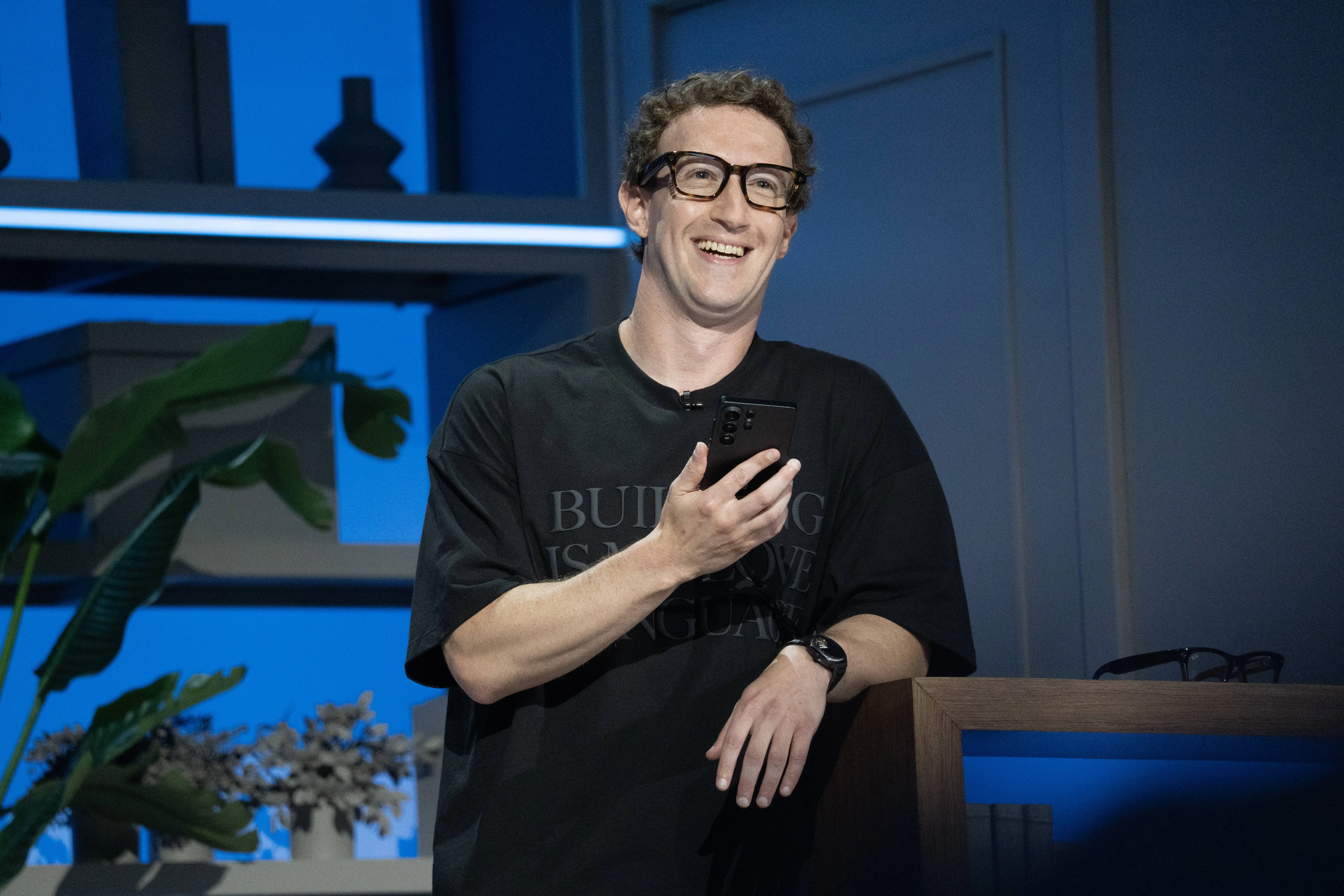 Mark Zuckerberg smiles while holding a smartphone on a blue-lit presentation stage.