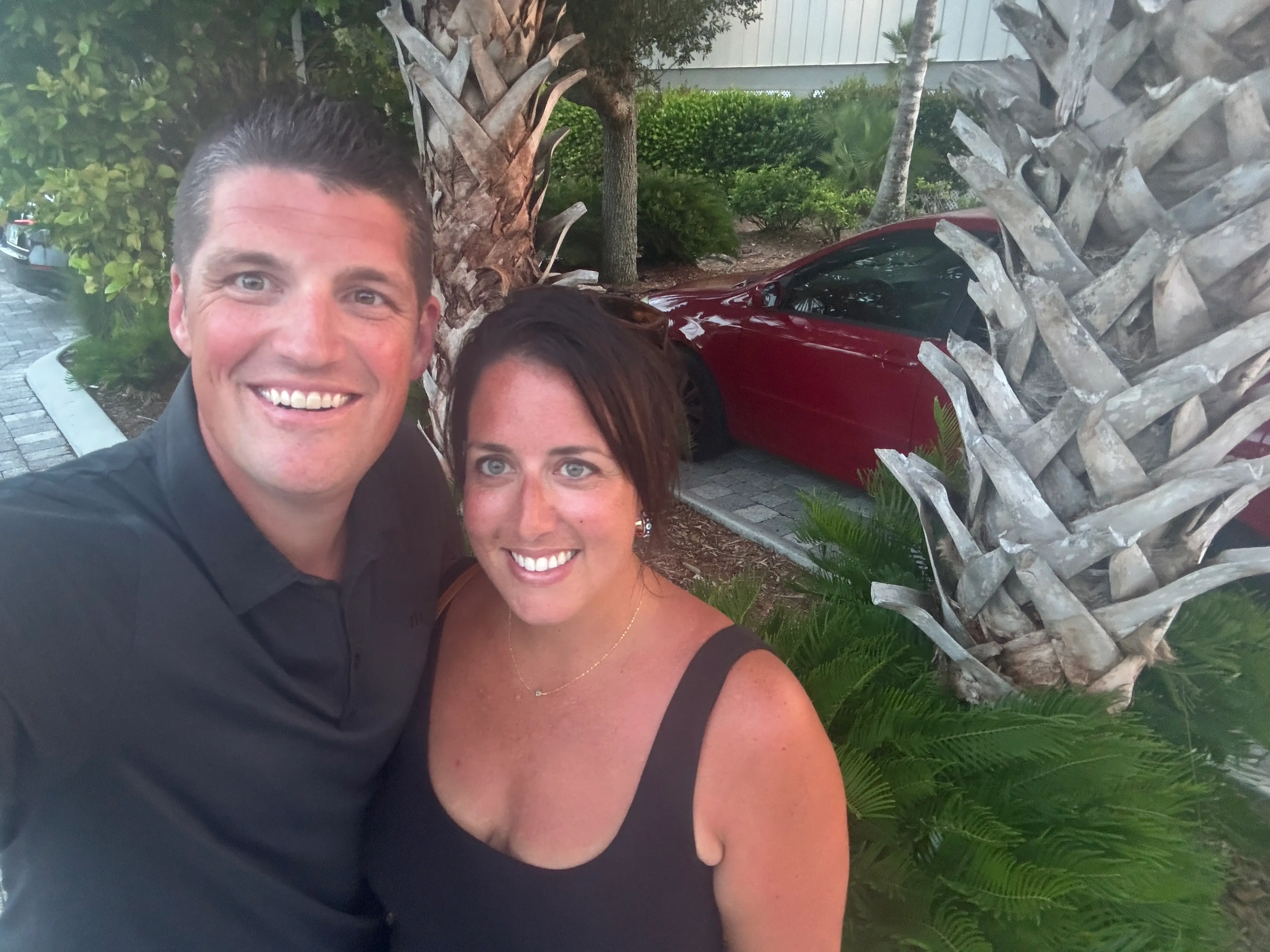 Man and woman smiling near palm trees, car, in selfie