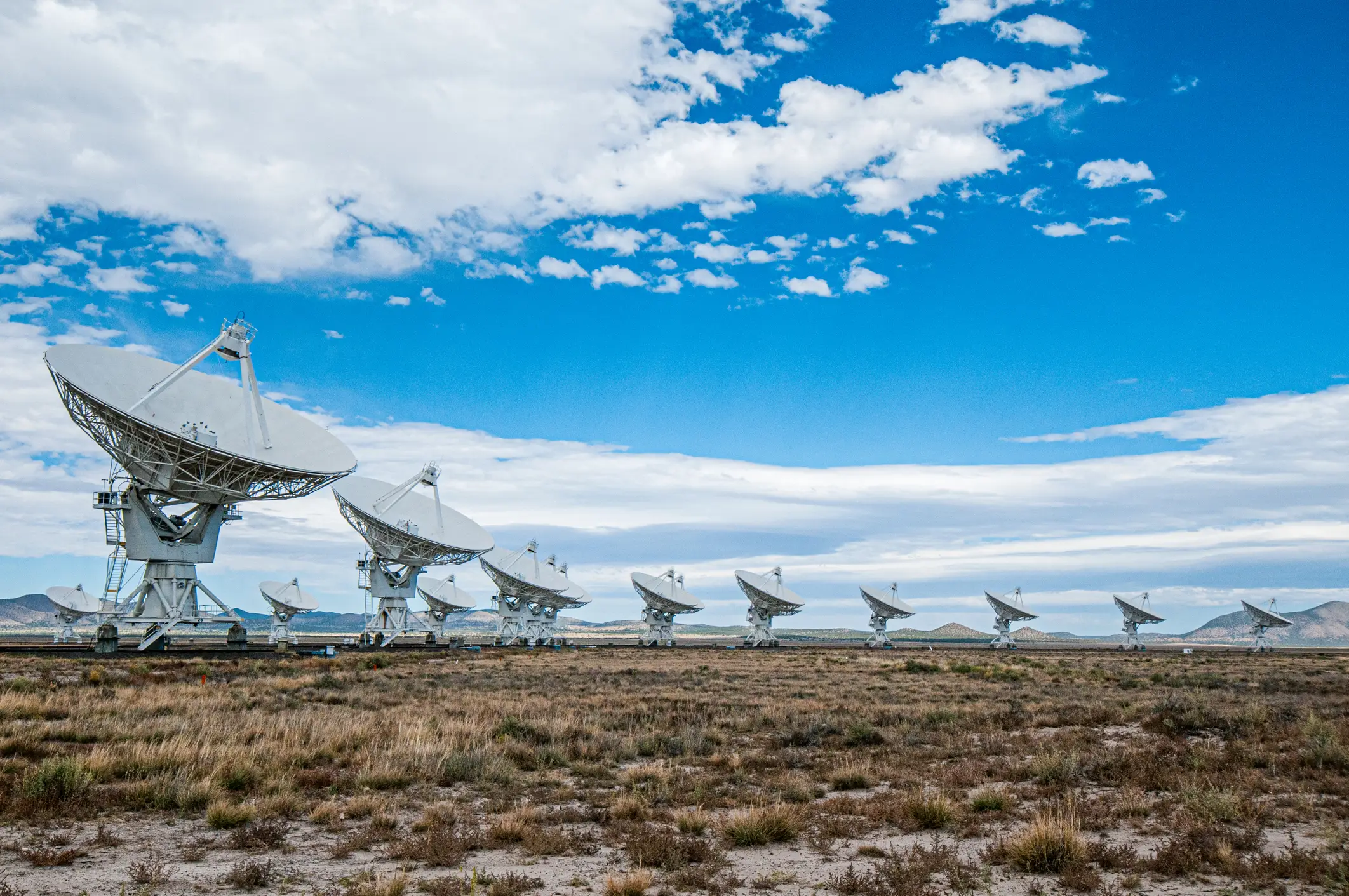 Radio telescope dishes line a desert plain beneath a blue sky with scattered clouds.