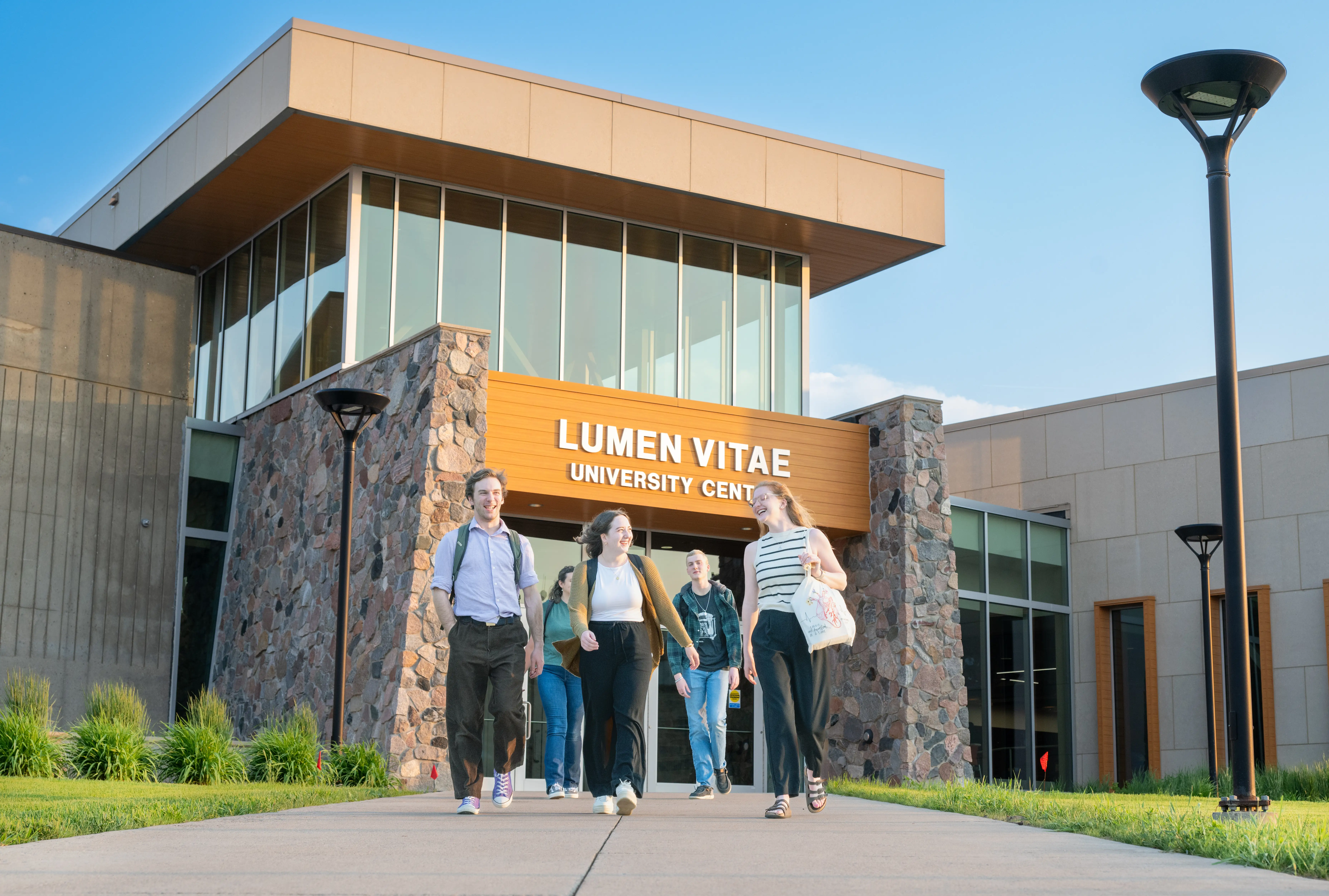 People with backpacks walk outside the LUMEN VITAE UNIVERSITY CENT entrance on a sunny campus walkway.
