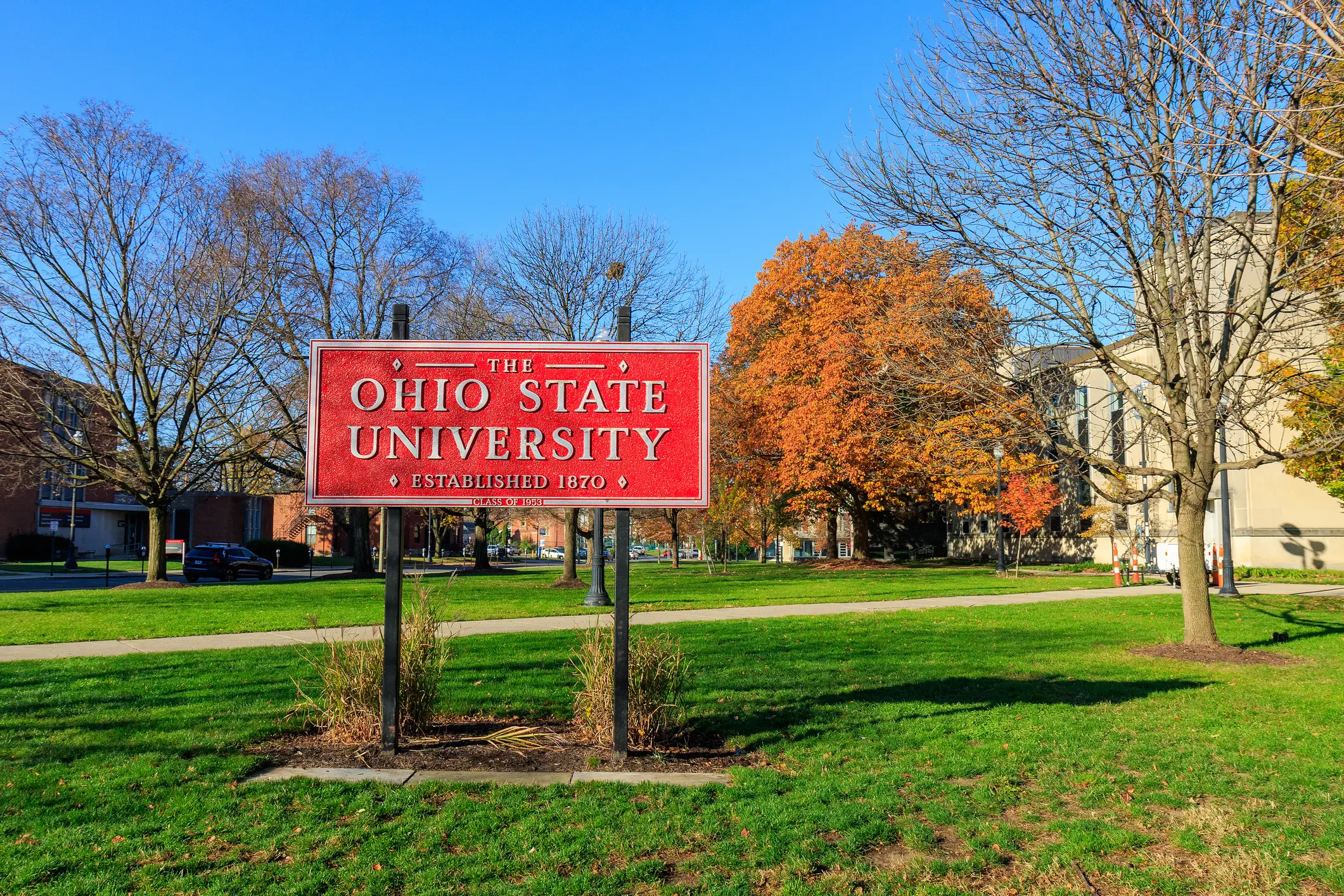 Red The Ohio State University sign stands on a grassy campus lawn with autumn trees and buildings.