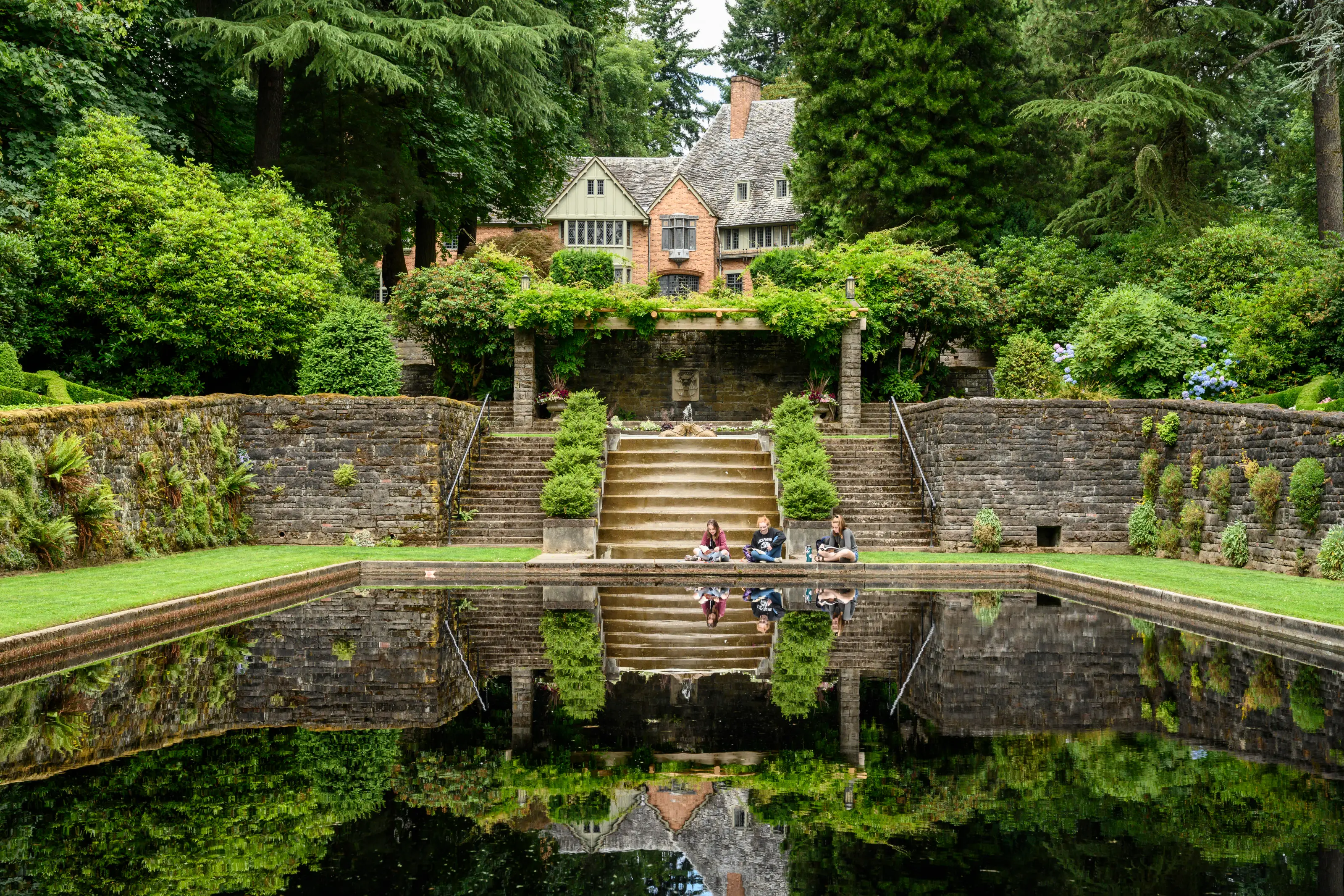 Three people sit beside a reflecting pool below stone terraces and a large house surrounded by lush gardens.