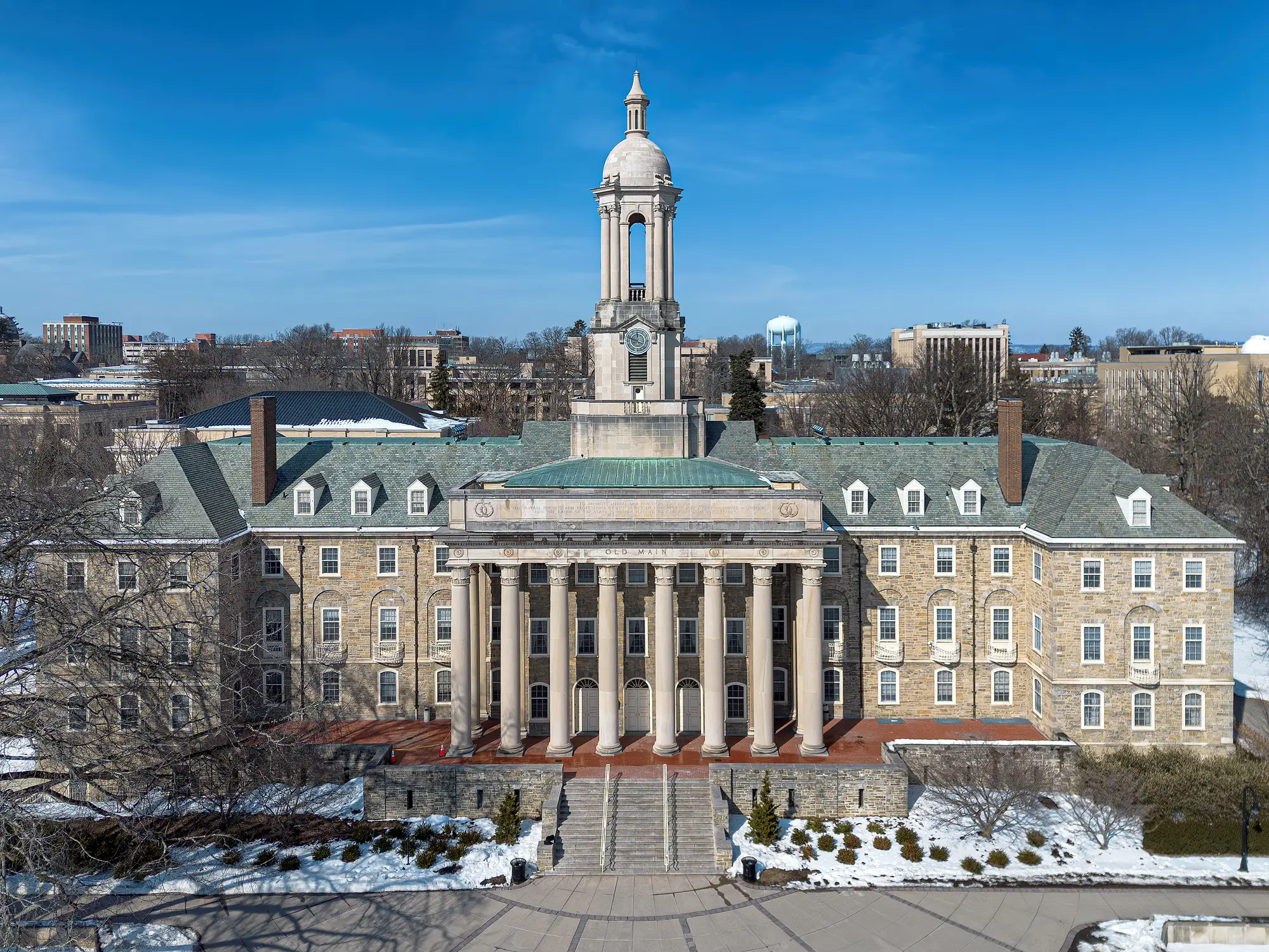 Old Main at Penn State stands behind snow-covered grounds under a clear blue sky.