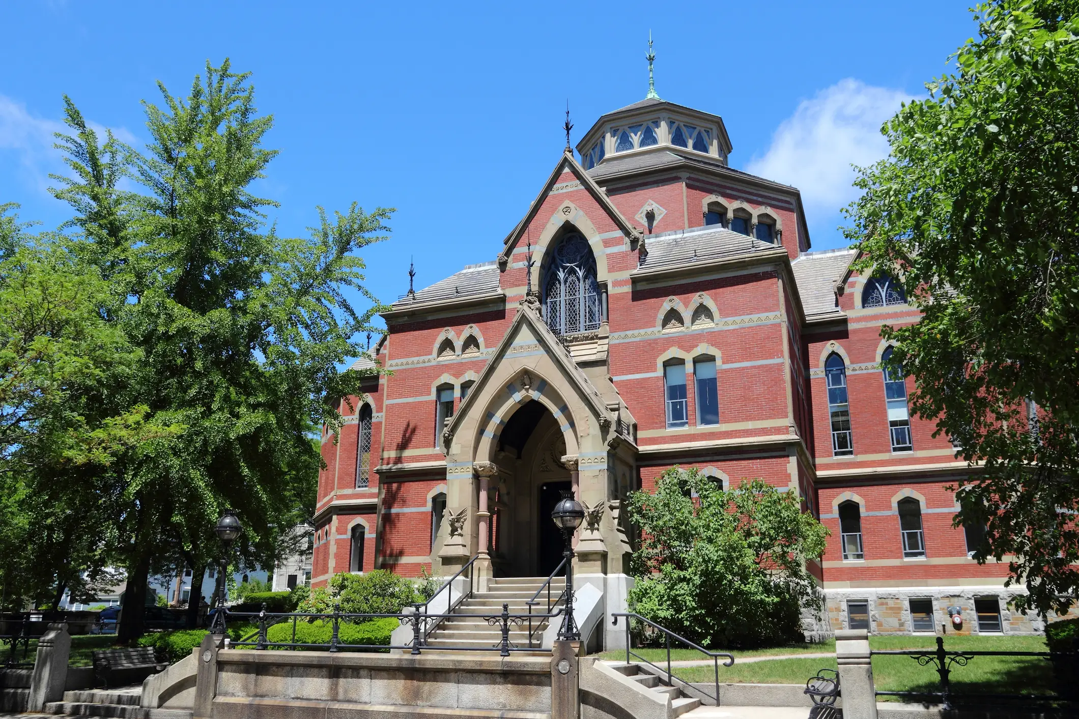 Red brick historic building with arched entrance, stairs, trees, and lampposts under a blue sky.