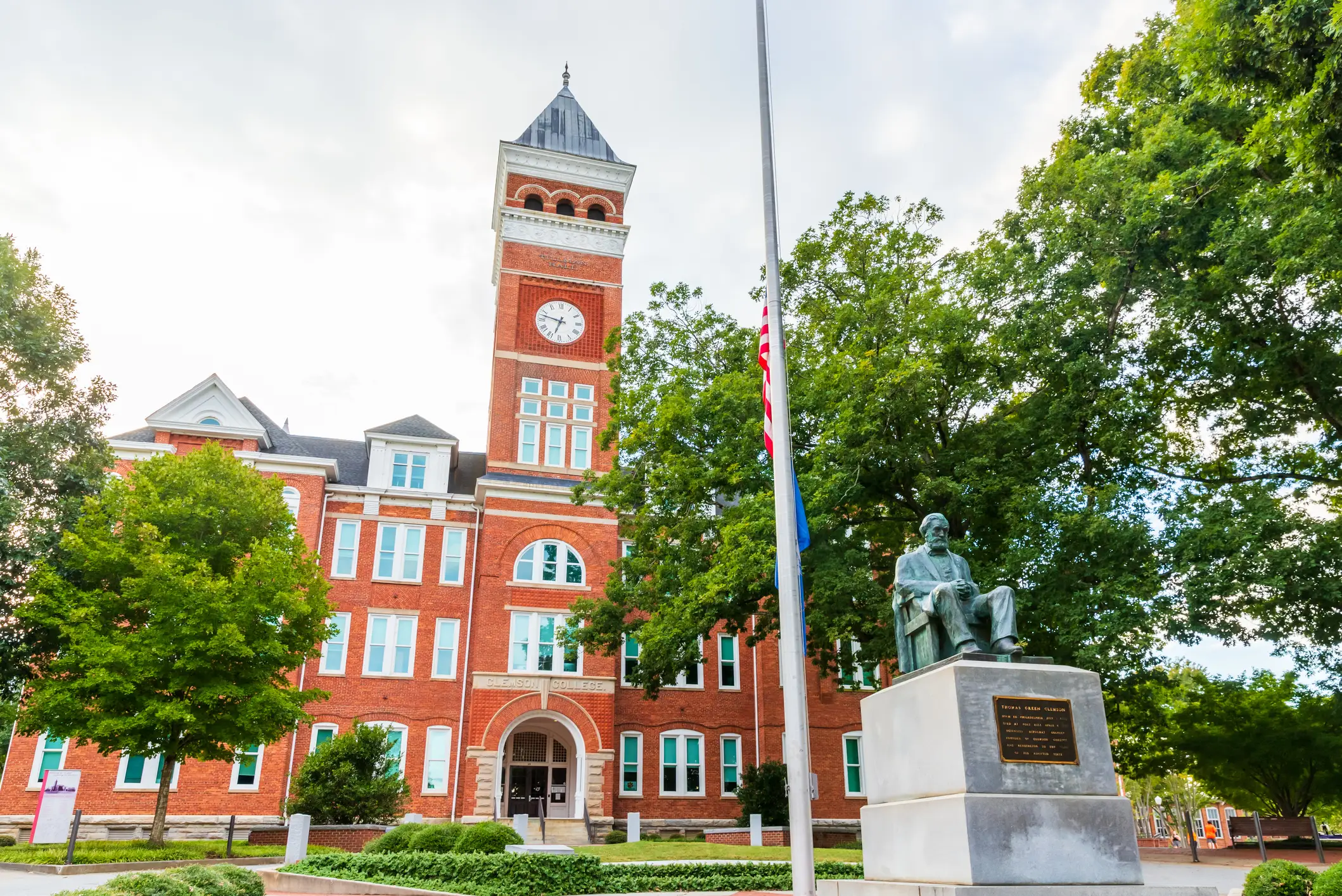 Clemson University’s red brick clock tower stands behind trees and a seated statue on landscaped campus grounds.