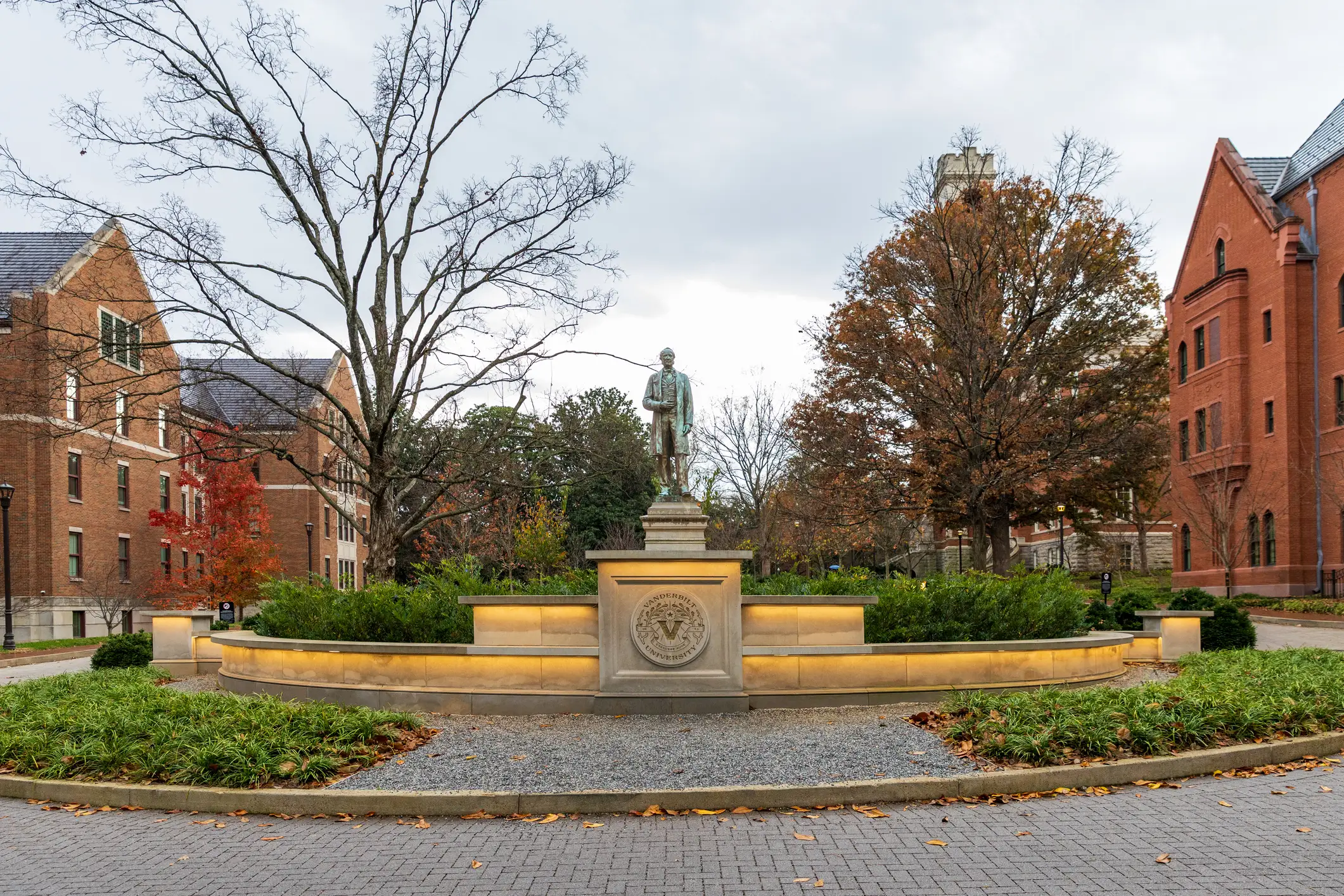 A statue and illuminated stone monument stand in a brick campus courtyard with autumn trees and red brick buildings.