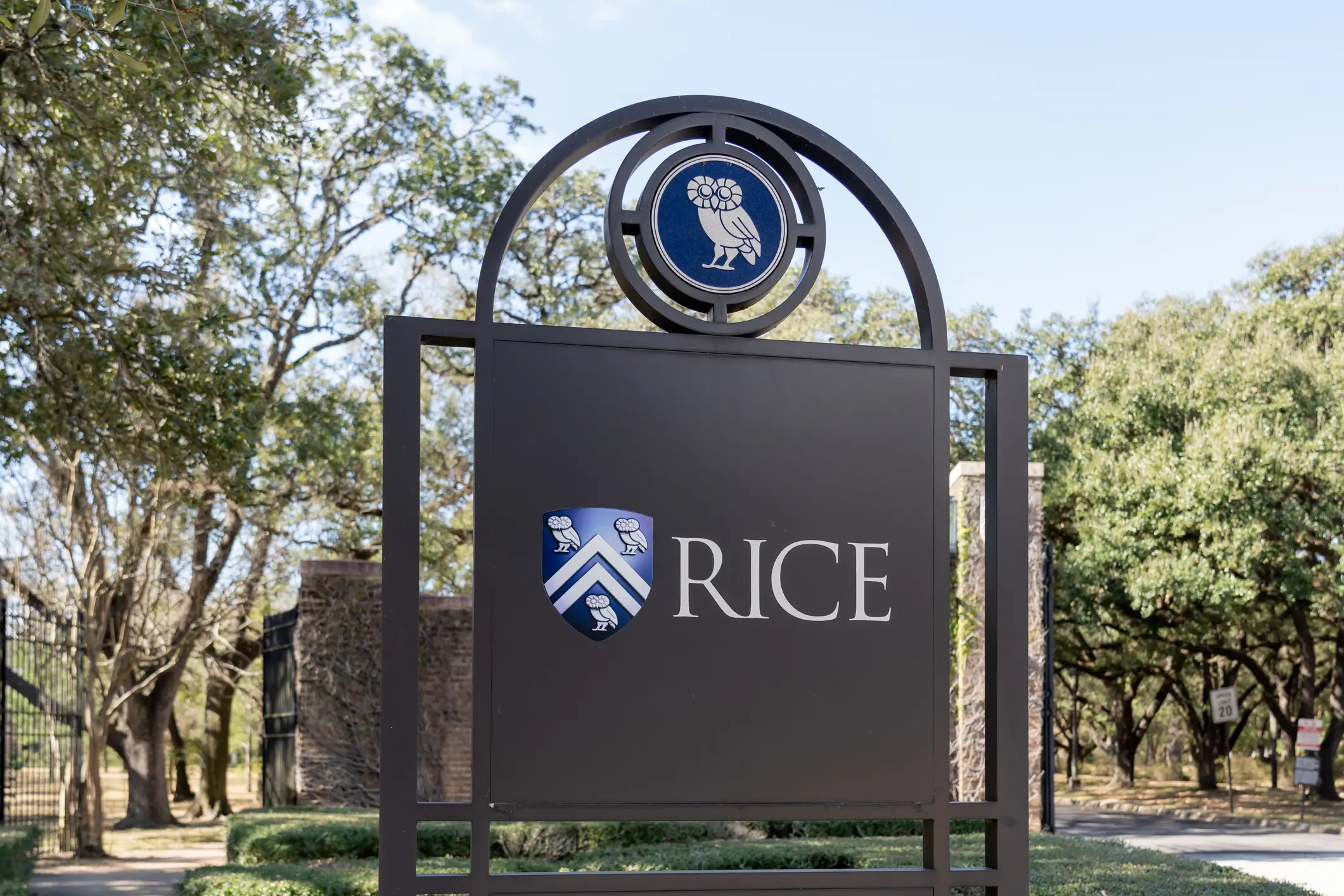 Rice University entrance sign with owl crest stands beside trees and a campus walkway.