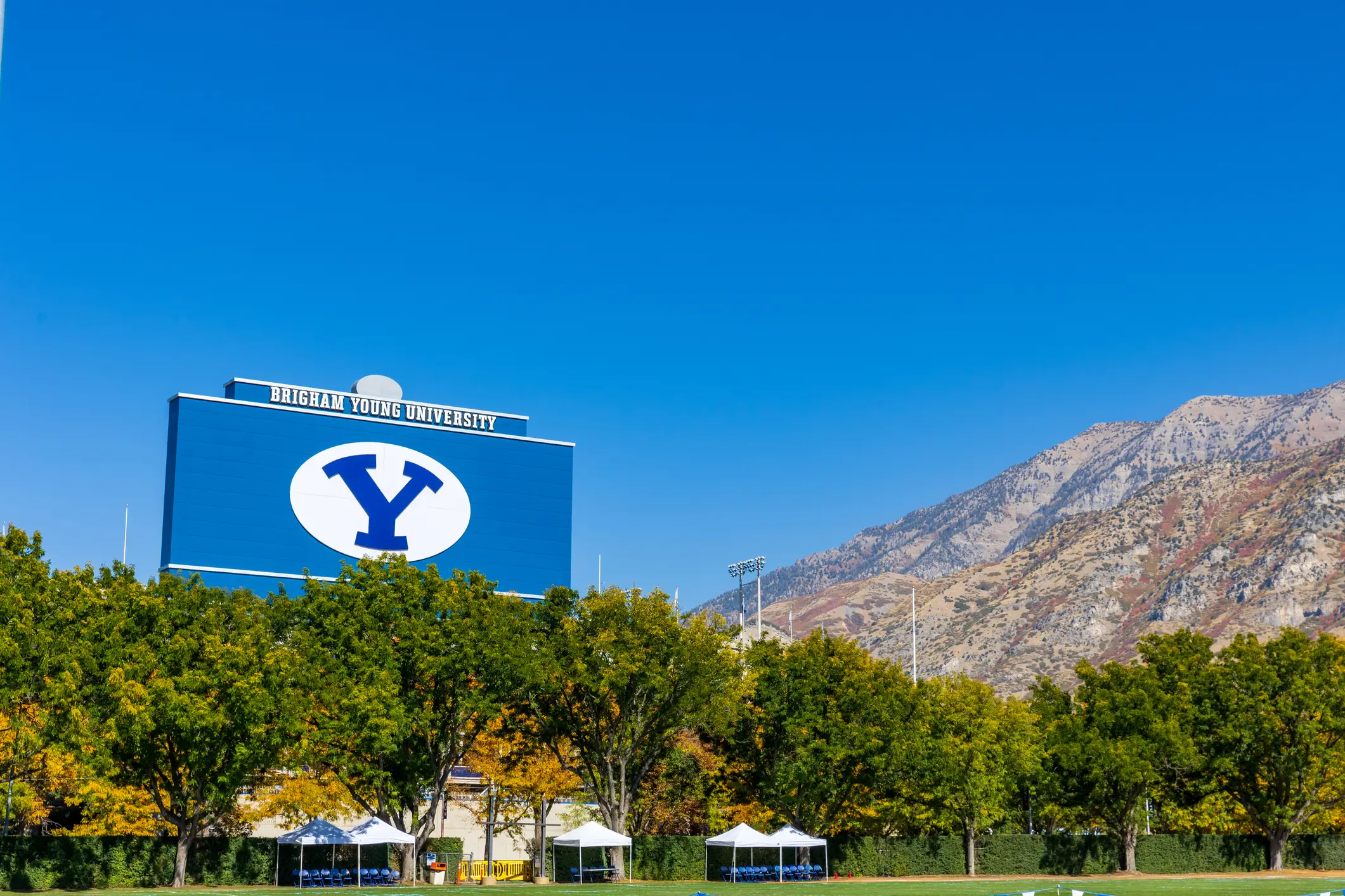 Brigham Young University scoreboard with a large Y logo stands beyond trees and a field below mountains.