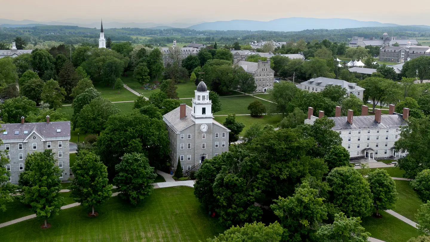 Aerial view of Middlebury College campus with Old Chapel, tree-lined lawns, stone buildings, and distant mountains.