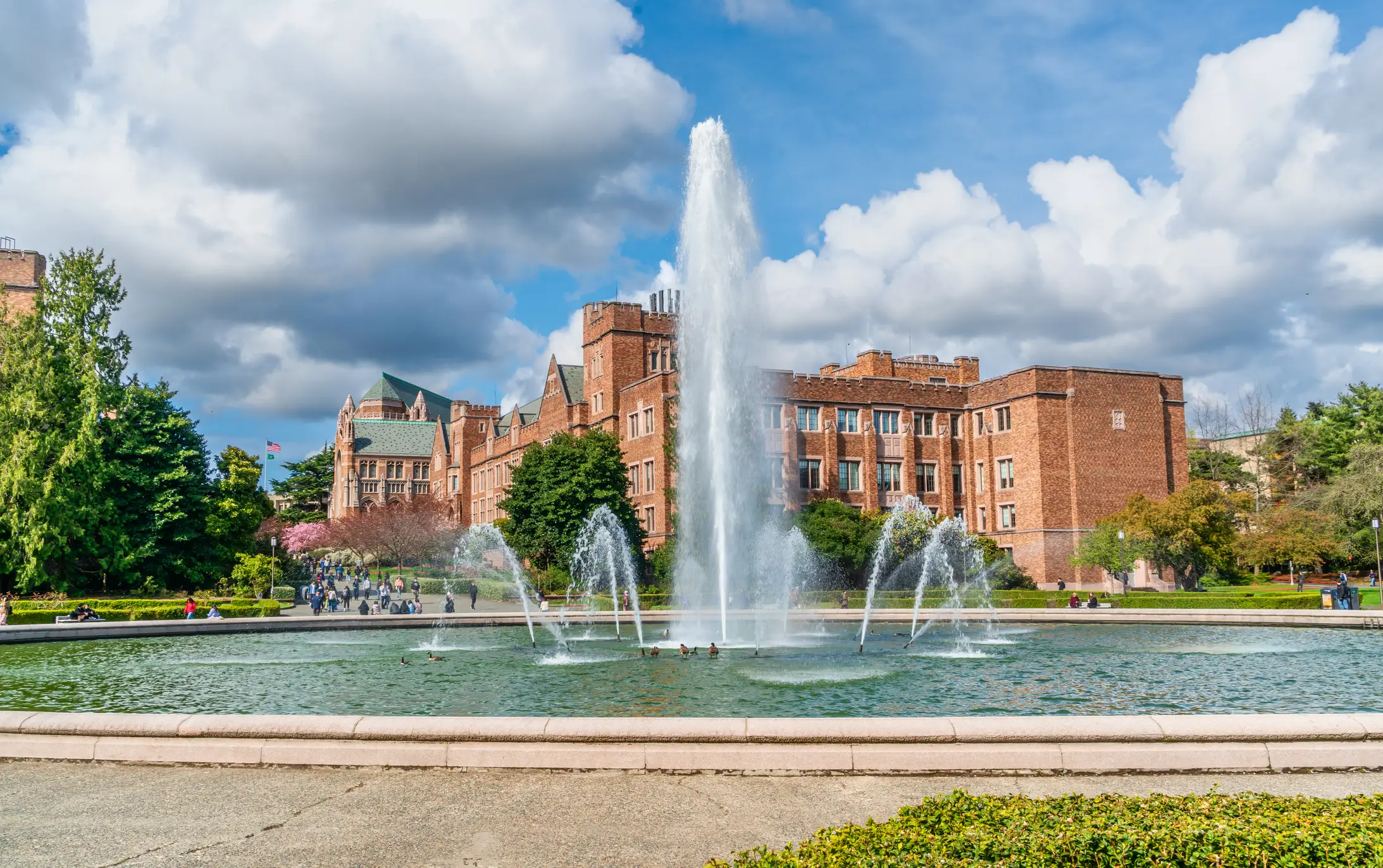Drumheller Fountain sprays in front of brick University of Washington campus buildings under a cloudy blue sky.