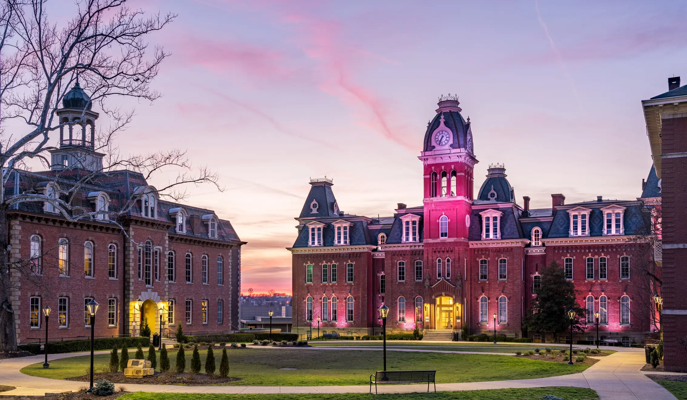 Red-brick university buildings with a lit clock tower surround a landscaped courtyard at sunset.