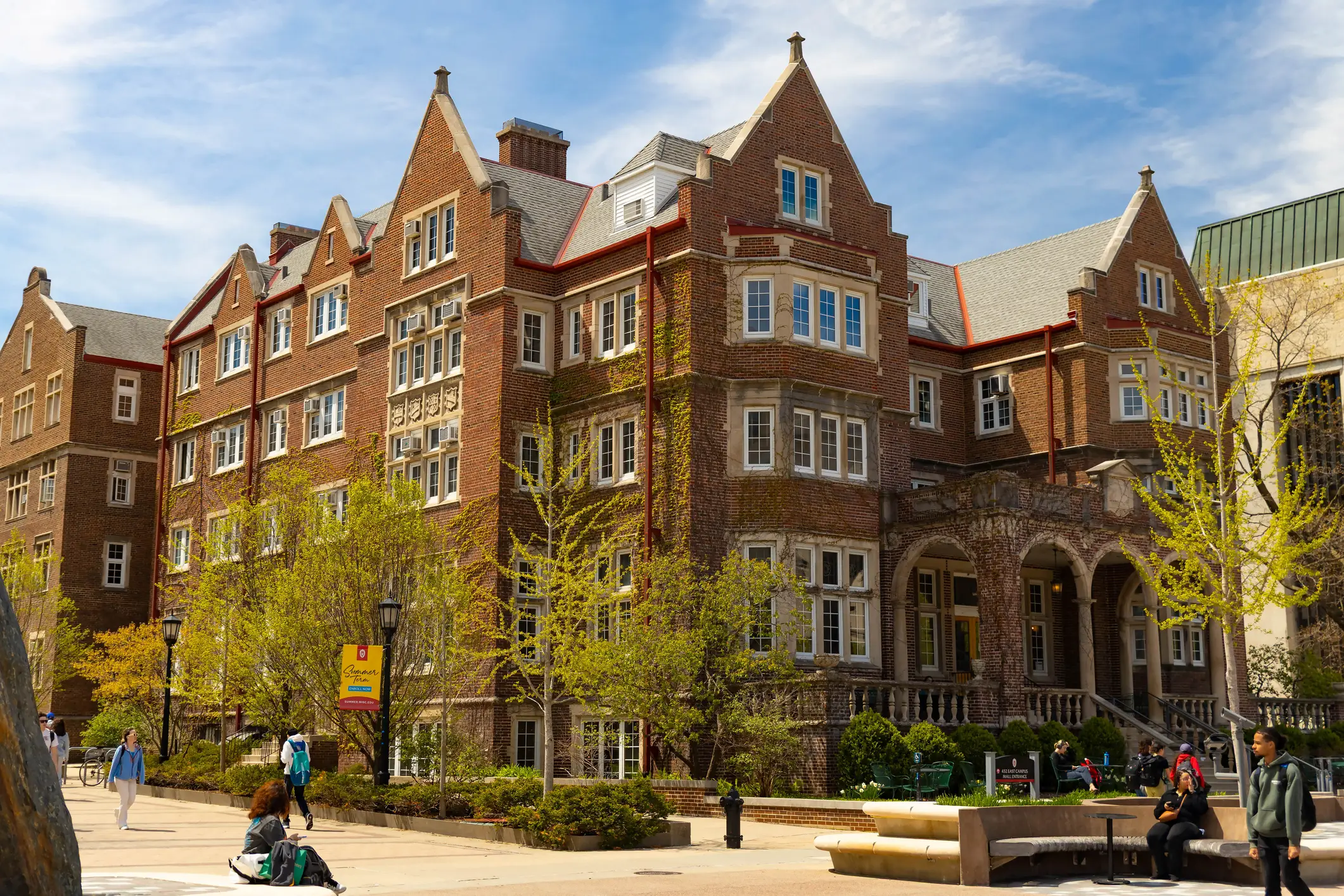 Red brick collegiate building with arched entrance and pedestrians on a sunny tree-lined campus walkway.