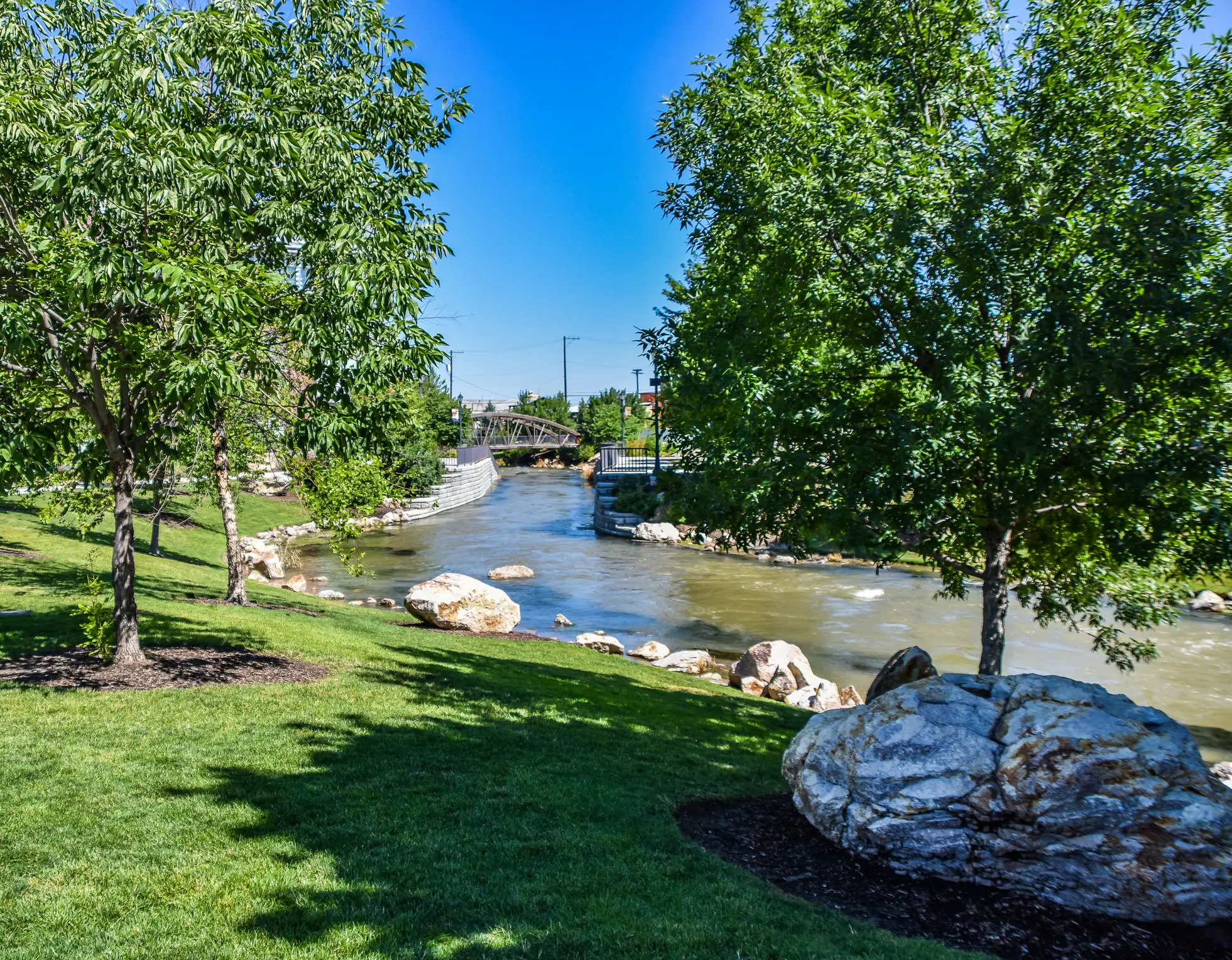 Tree-lined park with a river, large rocks, manicured grass, and a small bridge under a clear blue sky.