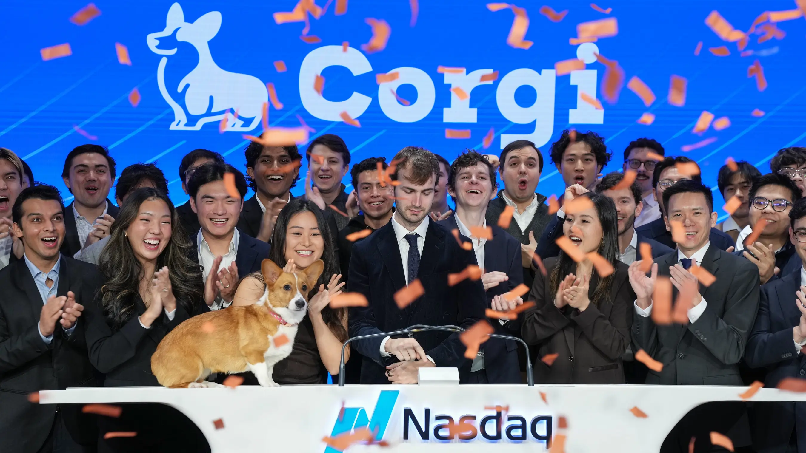 Group applauds around a corgi on a Nasdaq podium as orange confetti falls before a large Corgi screen.