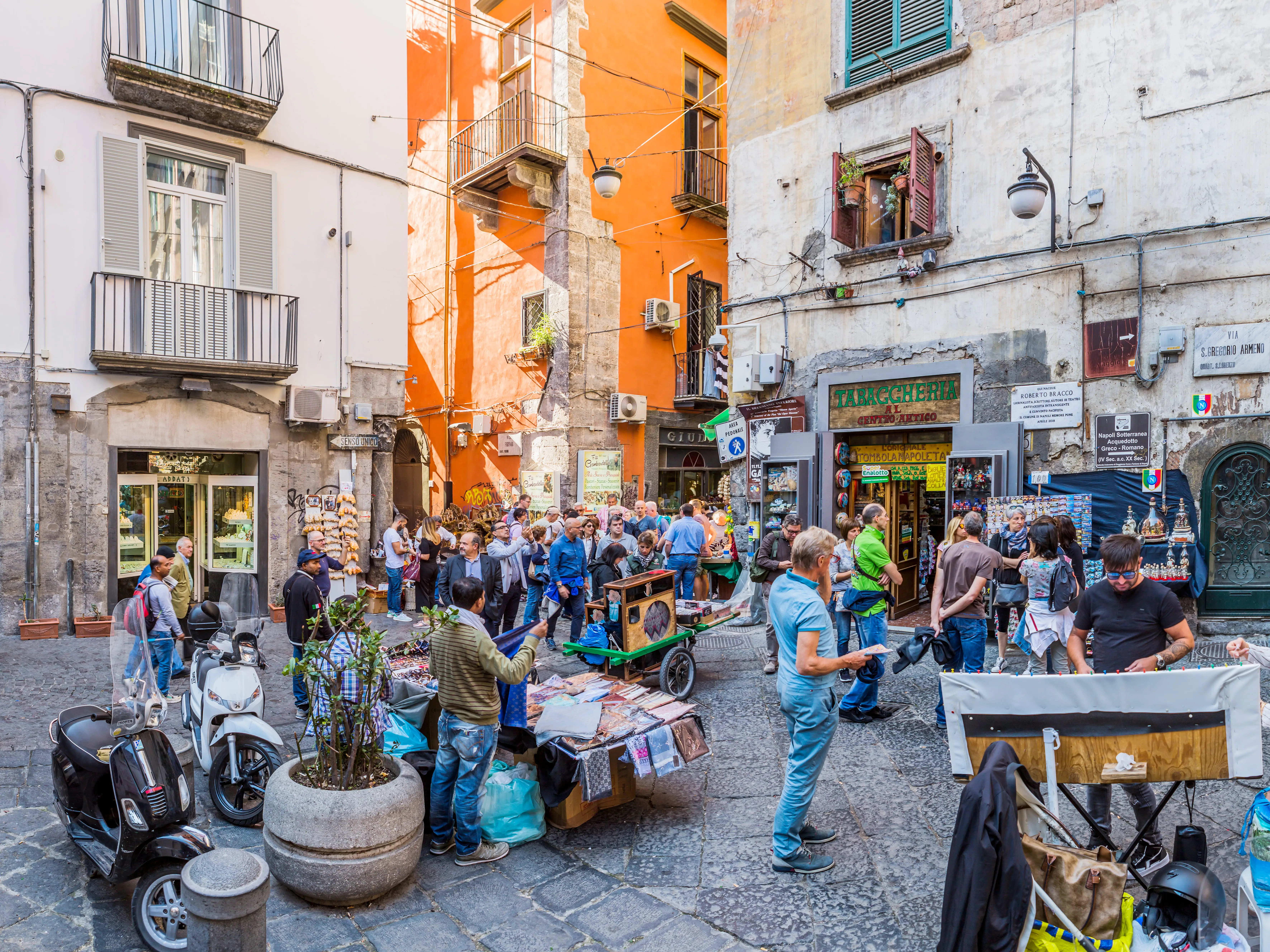 Crowds gather with scooters and street carts on a narrow urban street in Naples, Italy.