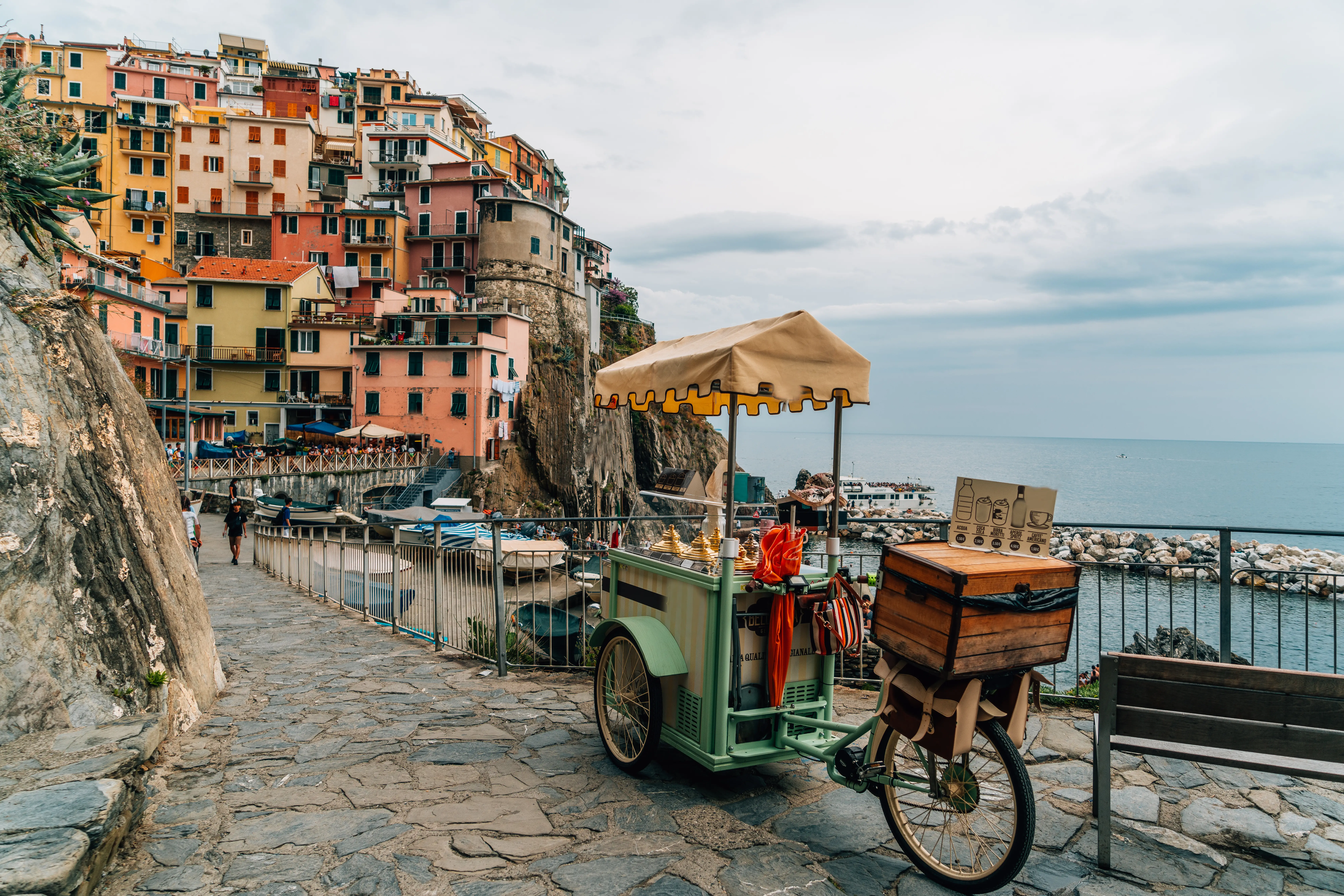 Colorful cliffside buildings overlook a stone seaside path with a vendor bicycle cart beside a harbor in Cinque Terre, Italy.