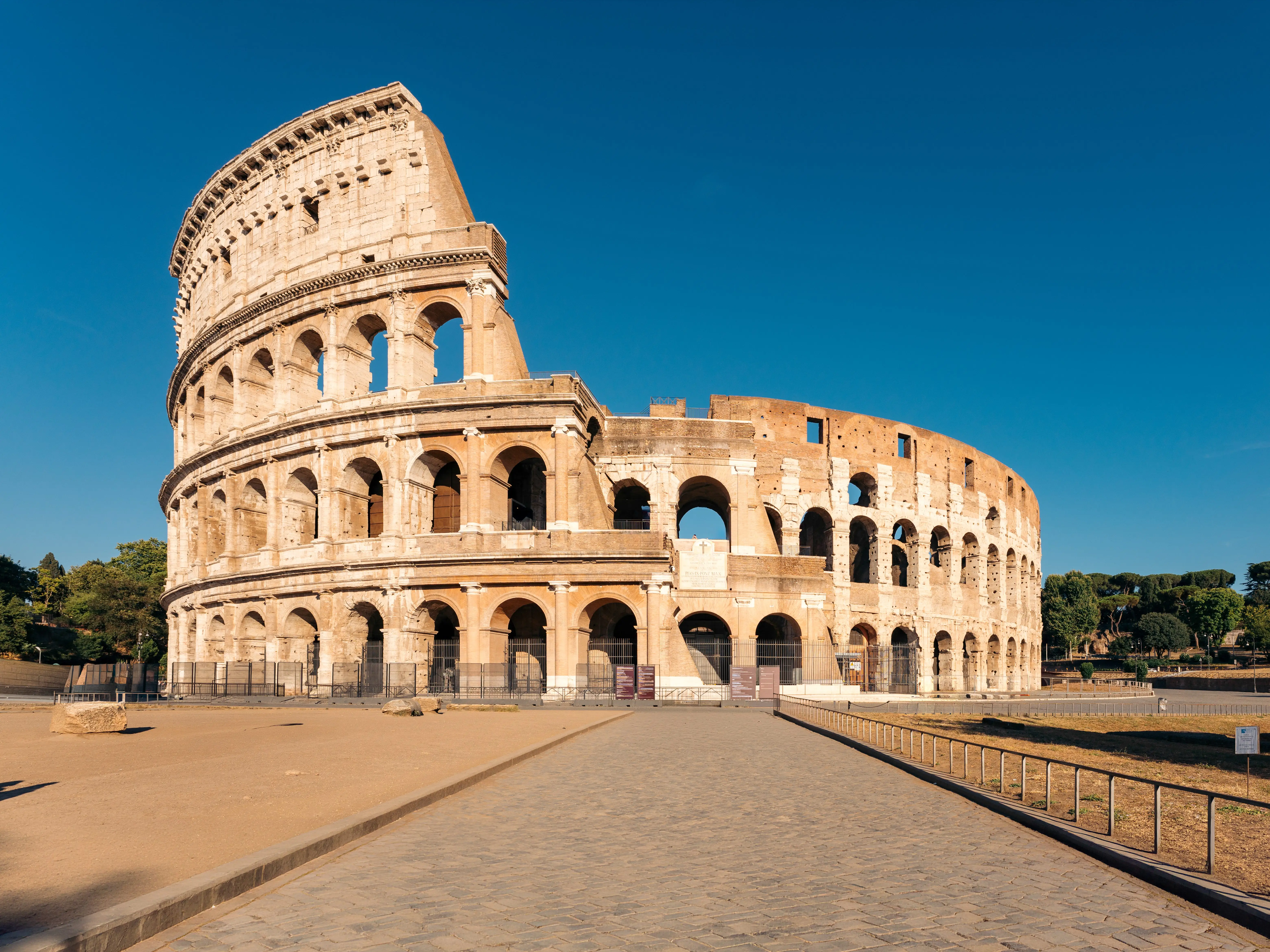 The Colosseum stands under a clear blue sky beside a wide paved plaza in Rome.