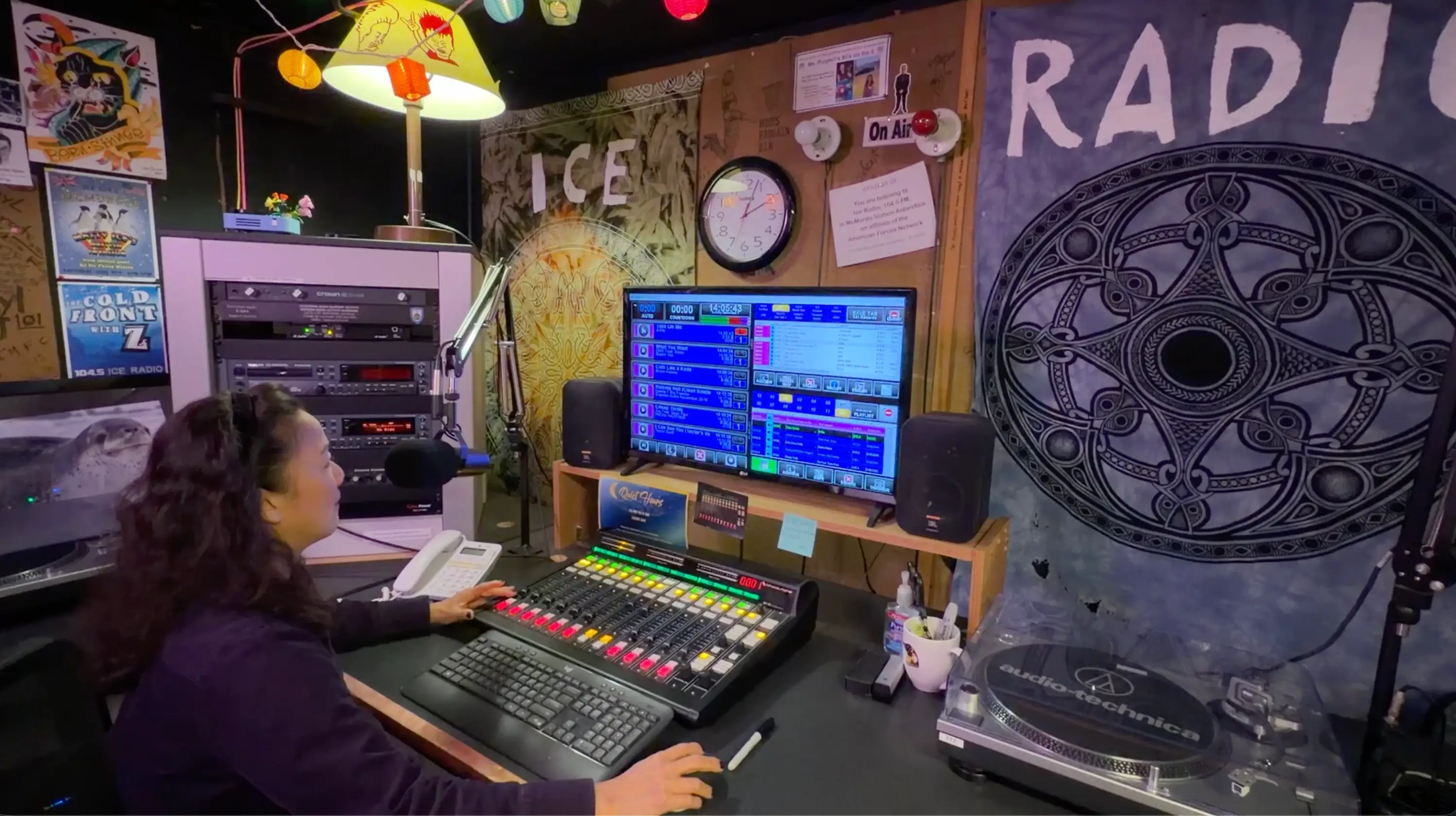 Person operates a mixing console and computer screens in a colorful radio studio with microphones and turntable.