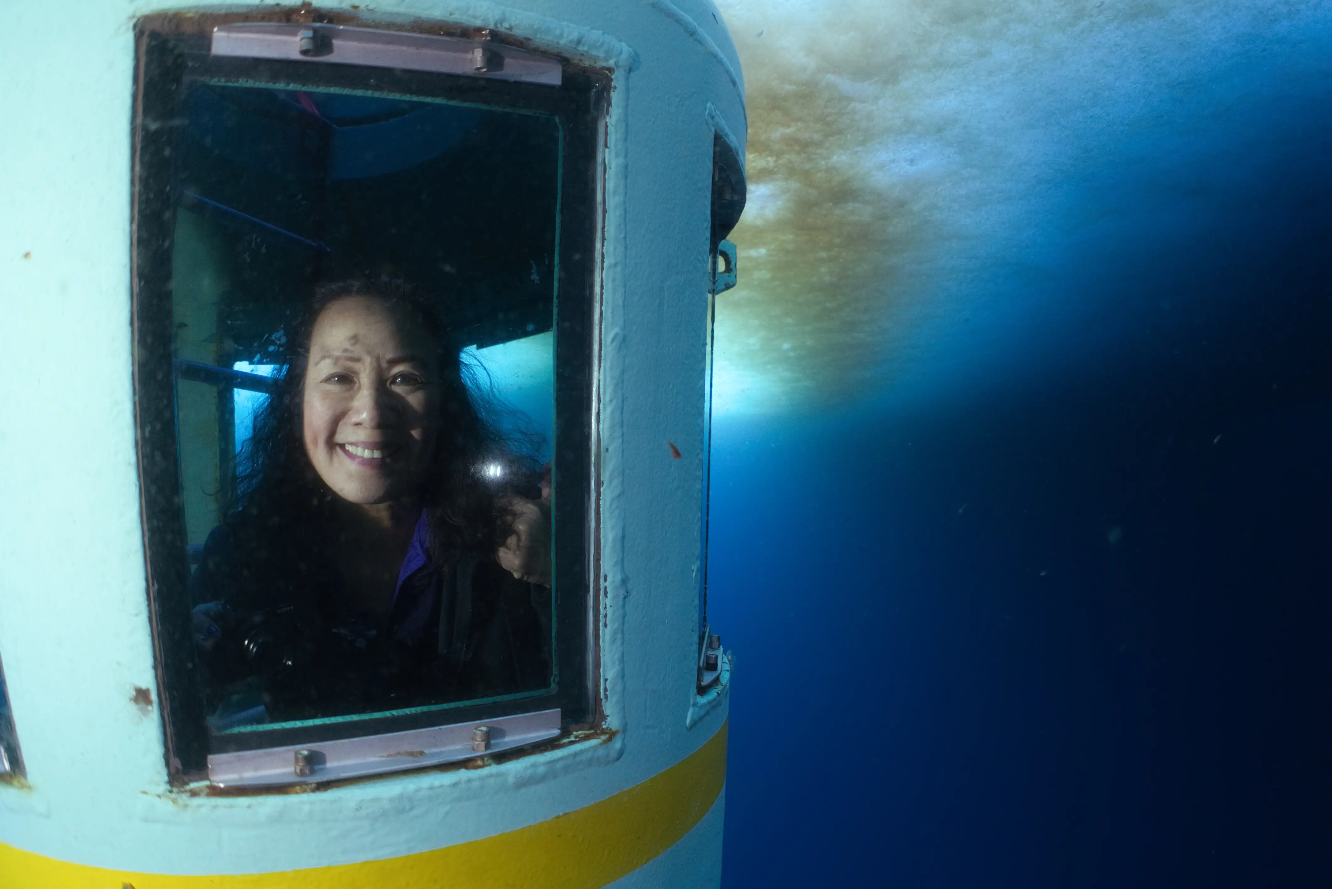 A smiling person looks through a window of an underwater vessel surrounded by deep blue water.