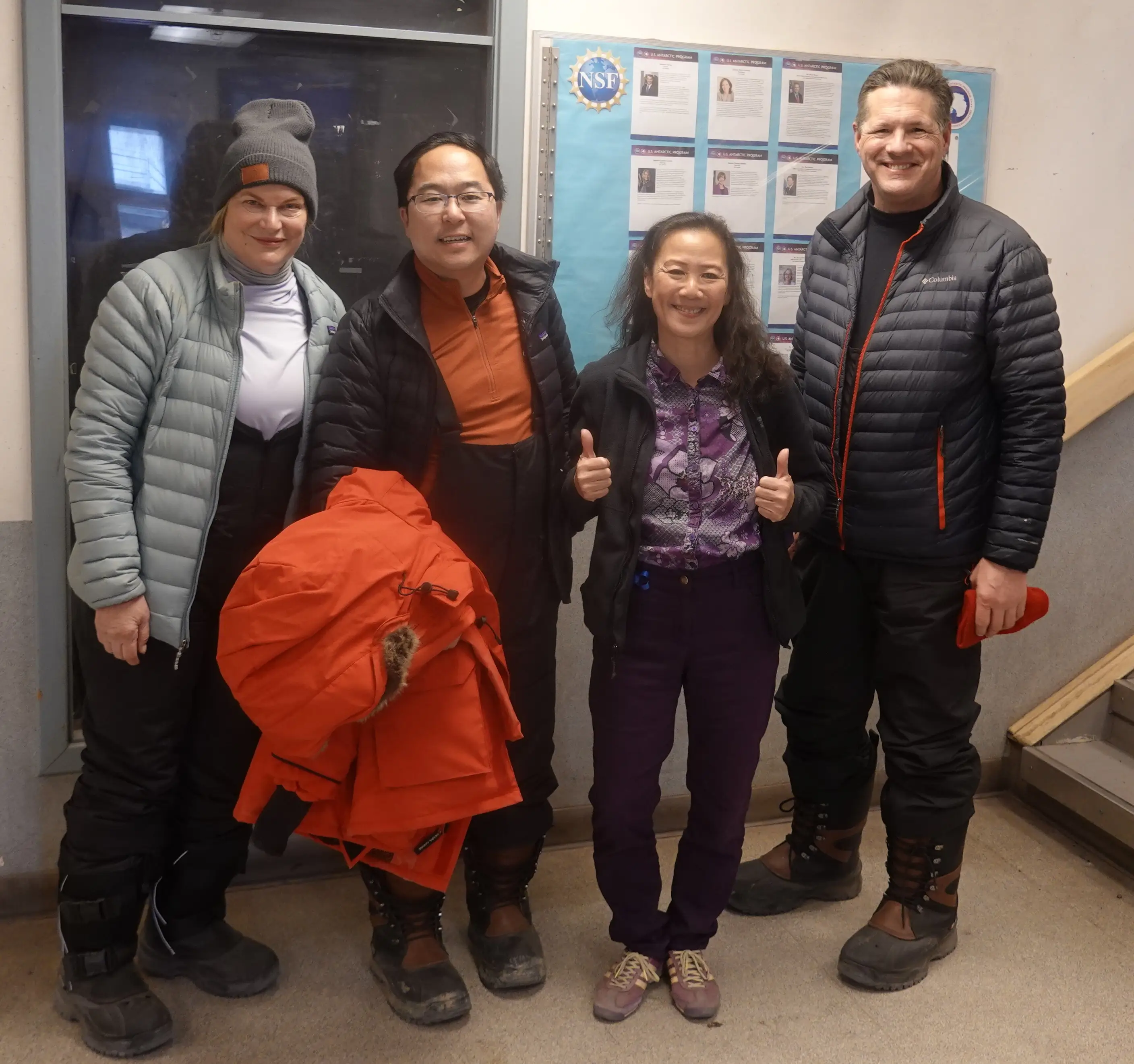 Four people in winter clothing pose indoors beside an NSF bulletin board, with one giving two thumbs up.