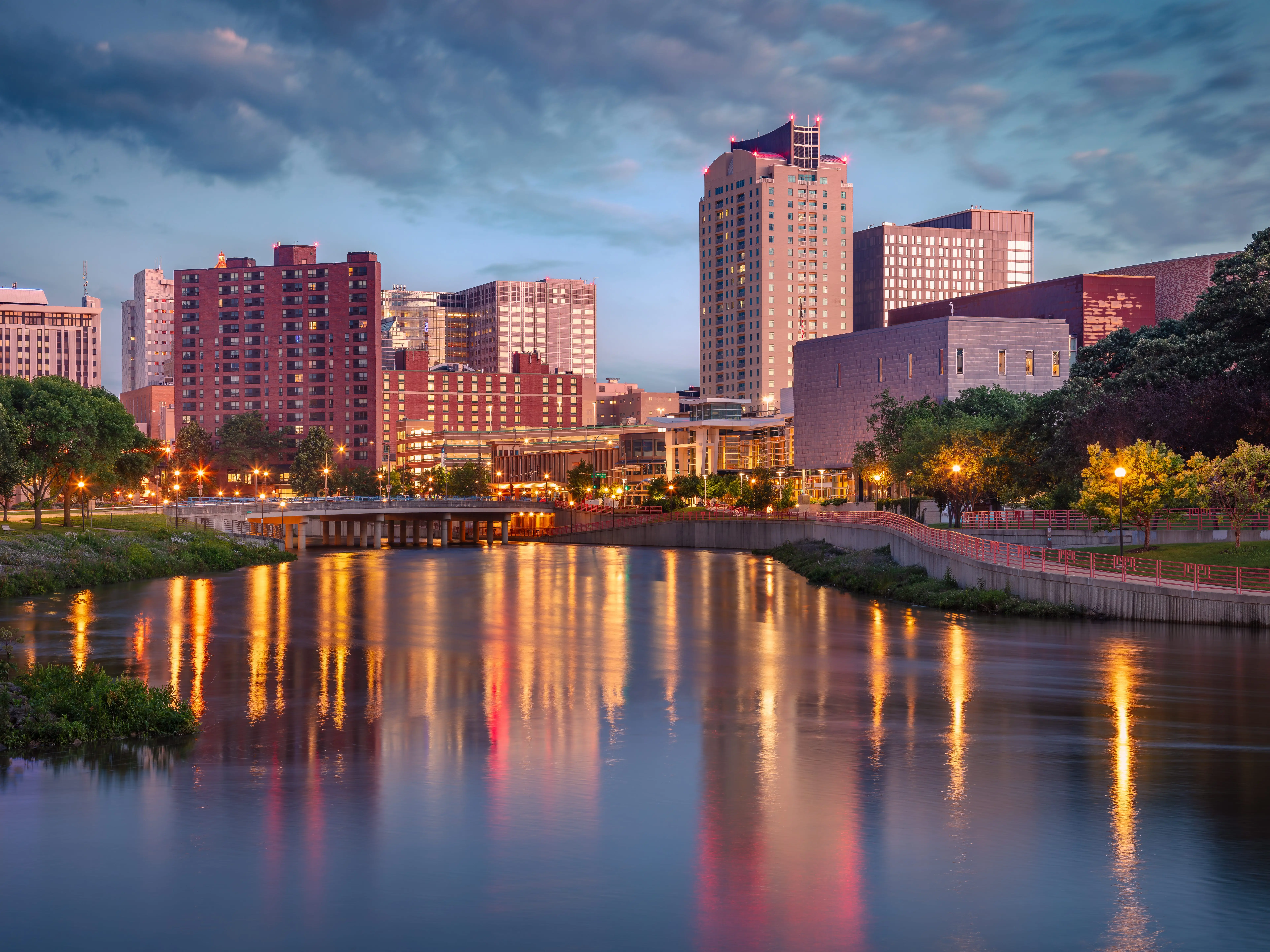 Rochester, Minnesota cityscape at summer sunrise features a river reflecting city lights. Skyscrapers and mid-rise buildings form the skyline, with varied architectural styles and heights.