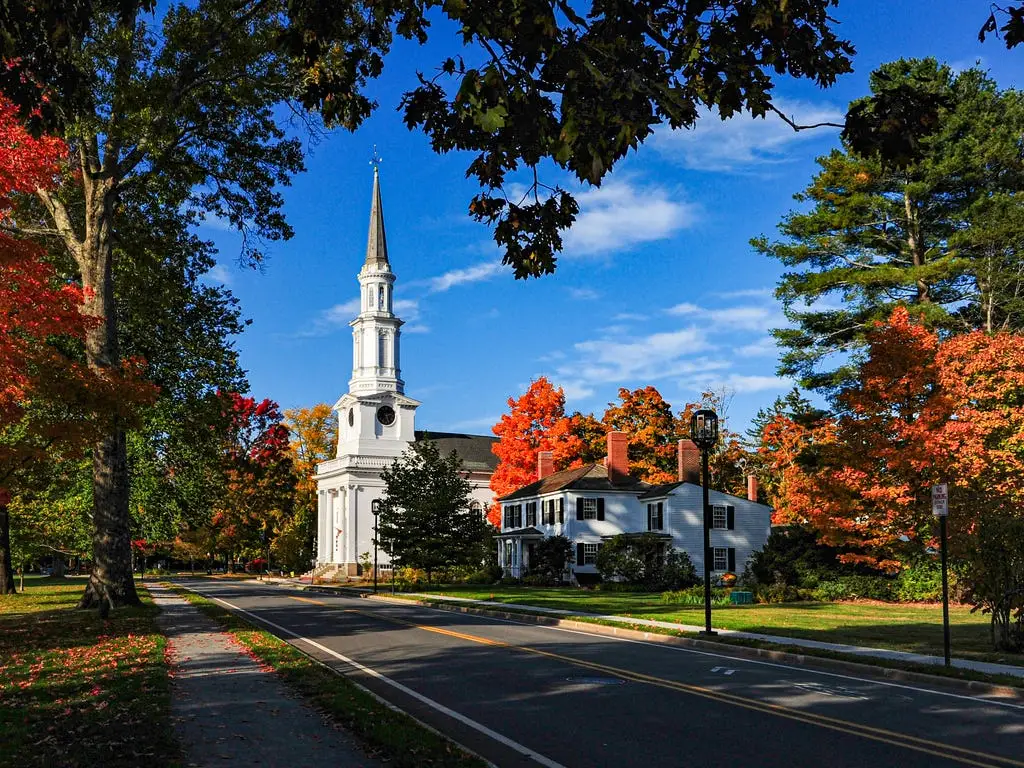 A white New England-style church with a tall steeple stands in a park covered with red and orange autumn leaves, surrounded by colorful trees under a bright blue sky.