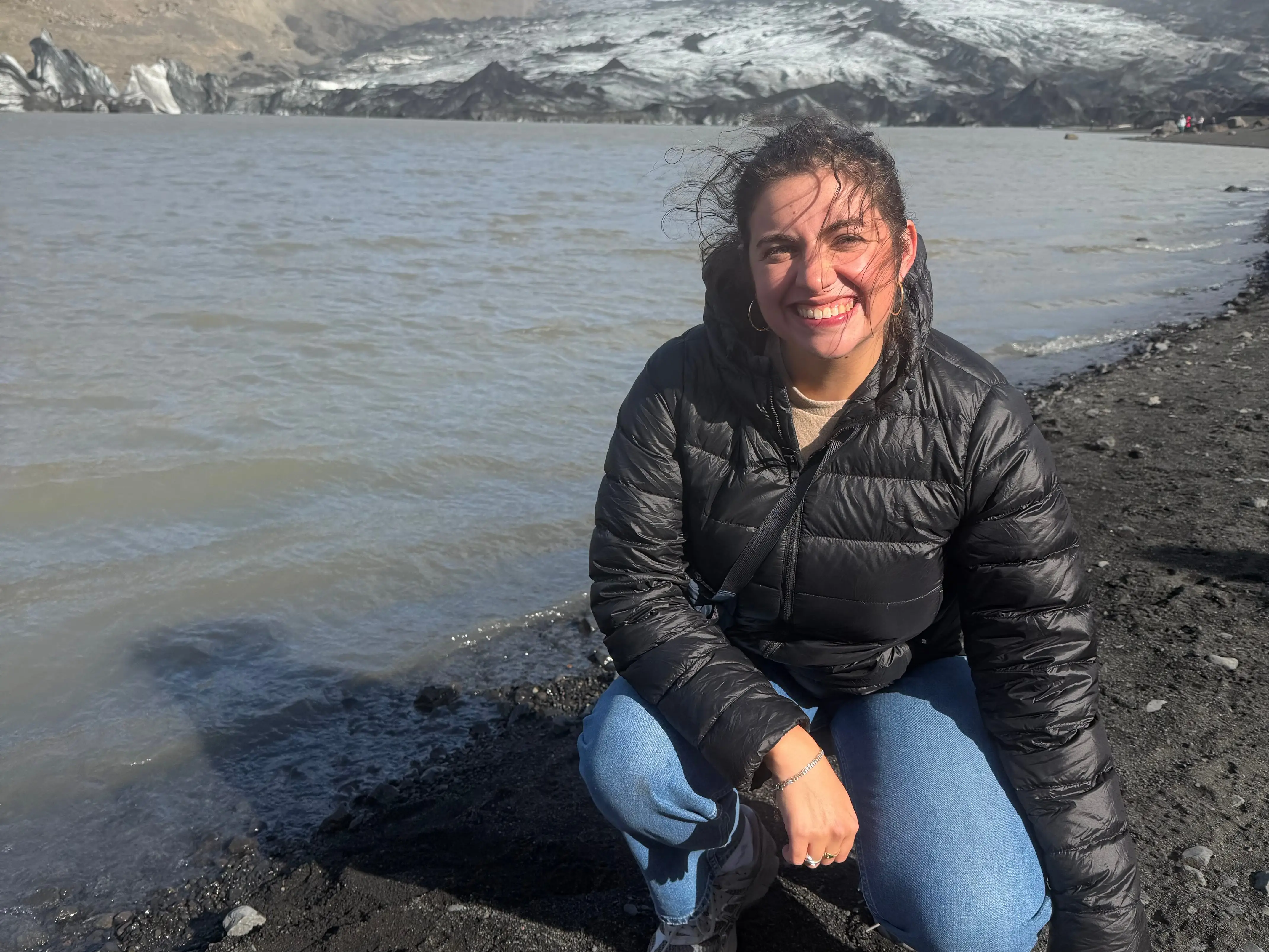 The writer crouches on a black-sand shoreline beside a glacial lagoon, with ice and mountains behind.