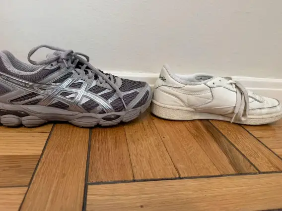 A gray Asics running shoe and a white Reebok sneaker sit side by side on a wooden floor against a wall.
