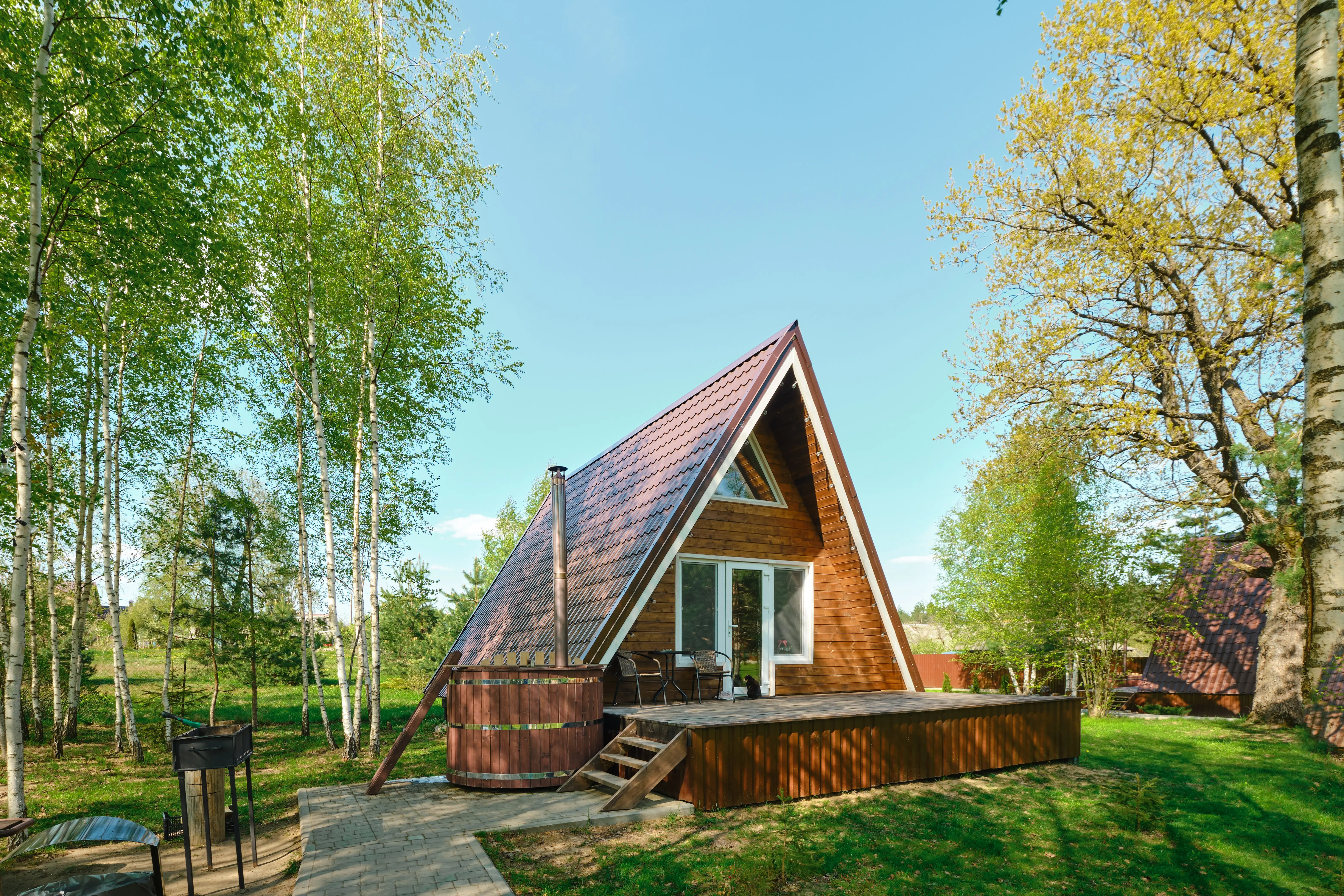 A-frame wooden cabin with a deck and hot tub sits in a sunny green forest clearing.