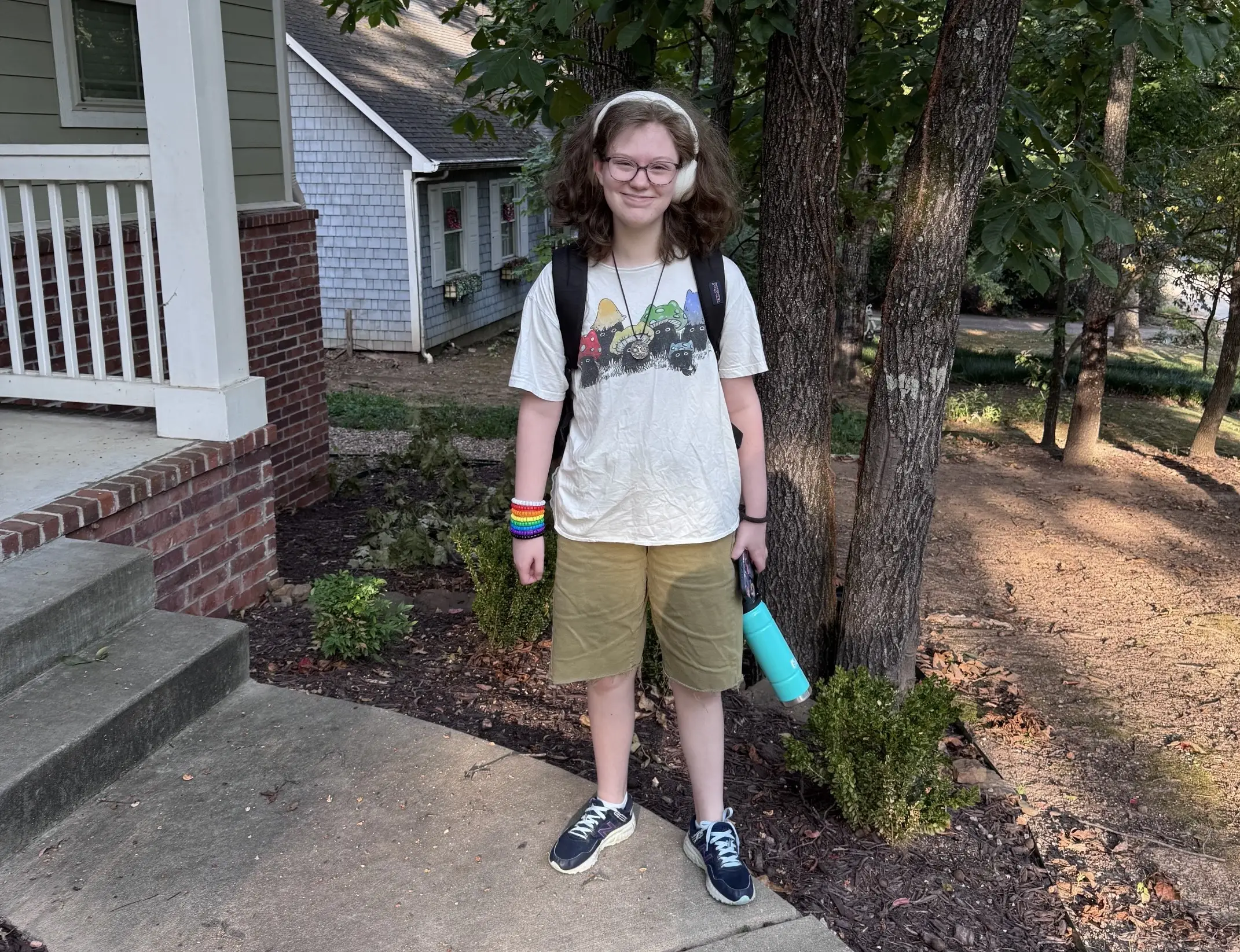 Person with backpack and headphones stands outside a house near trees holding a turquoise water bottle.