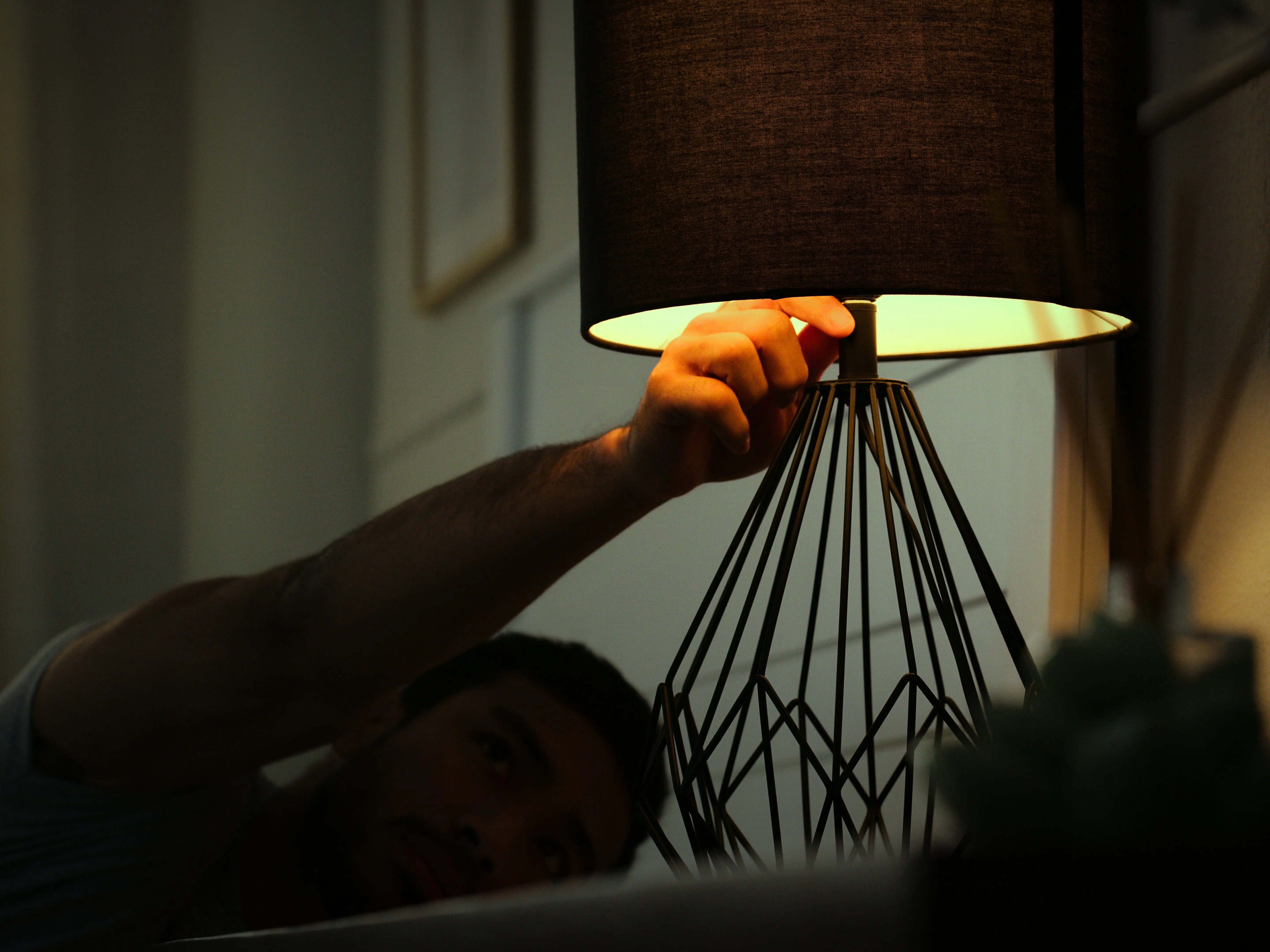 A partially obscured person reaches toward a lit table lamp in a dark interior.