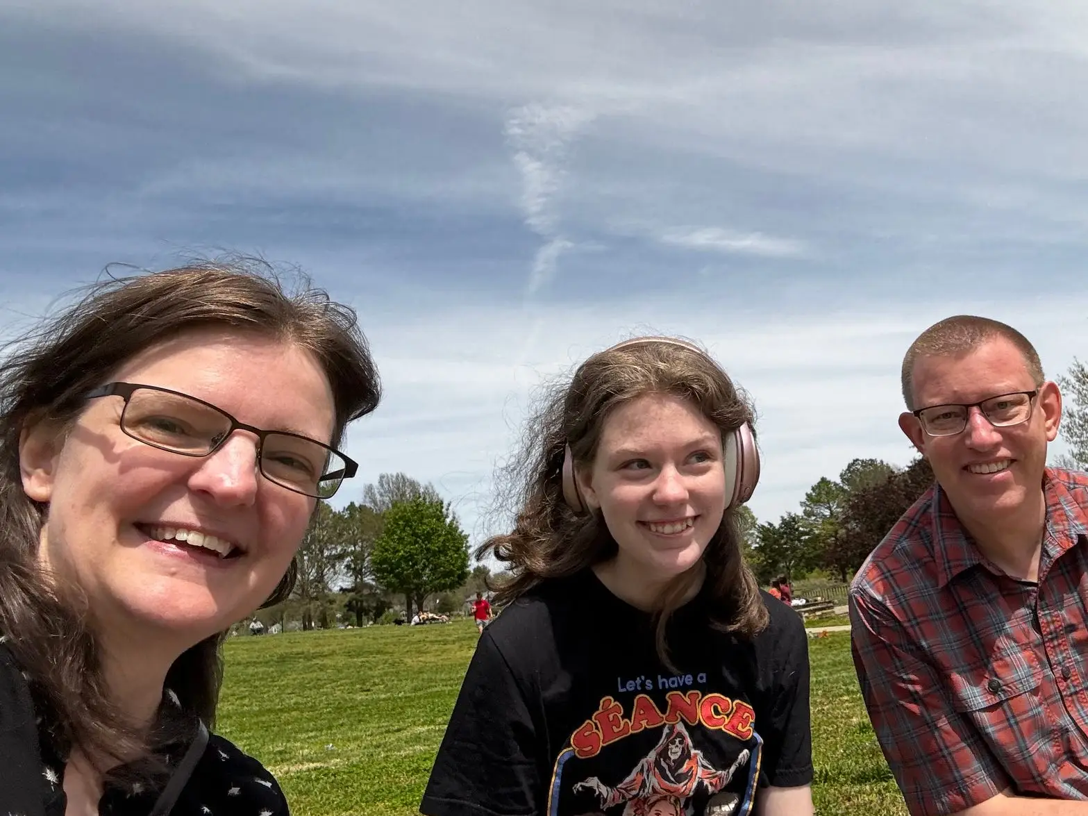 Three smiling people sit on grass in a park under a cloudy sky, with trees in the background.