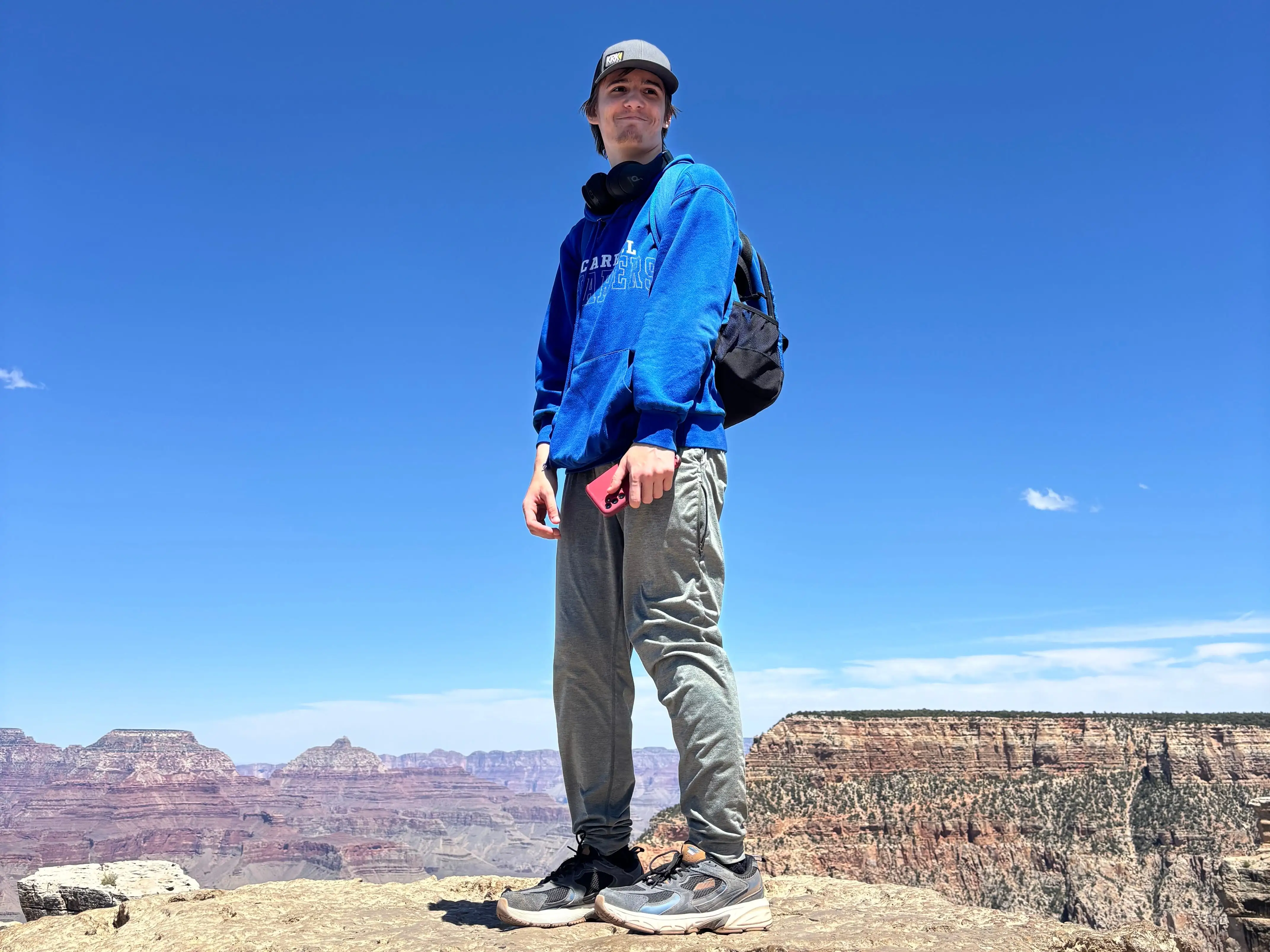 Person in blue hoodie stands on a rocky canyon overlook beneath a clear blue sky.
