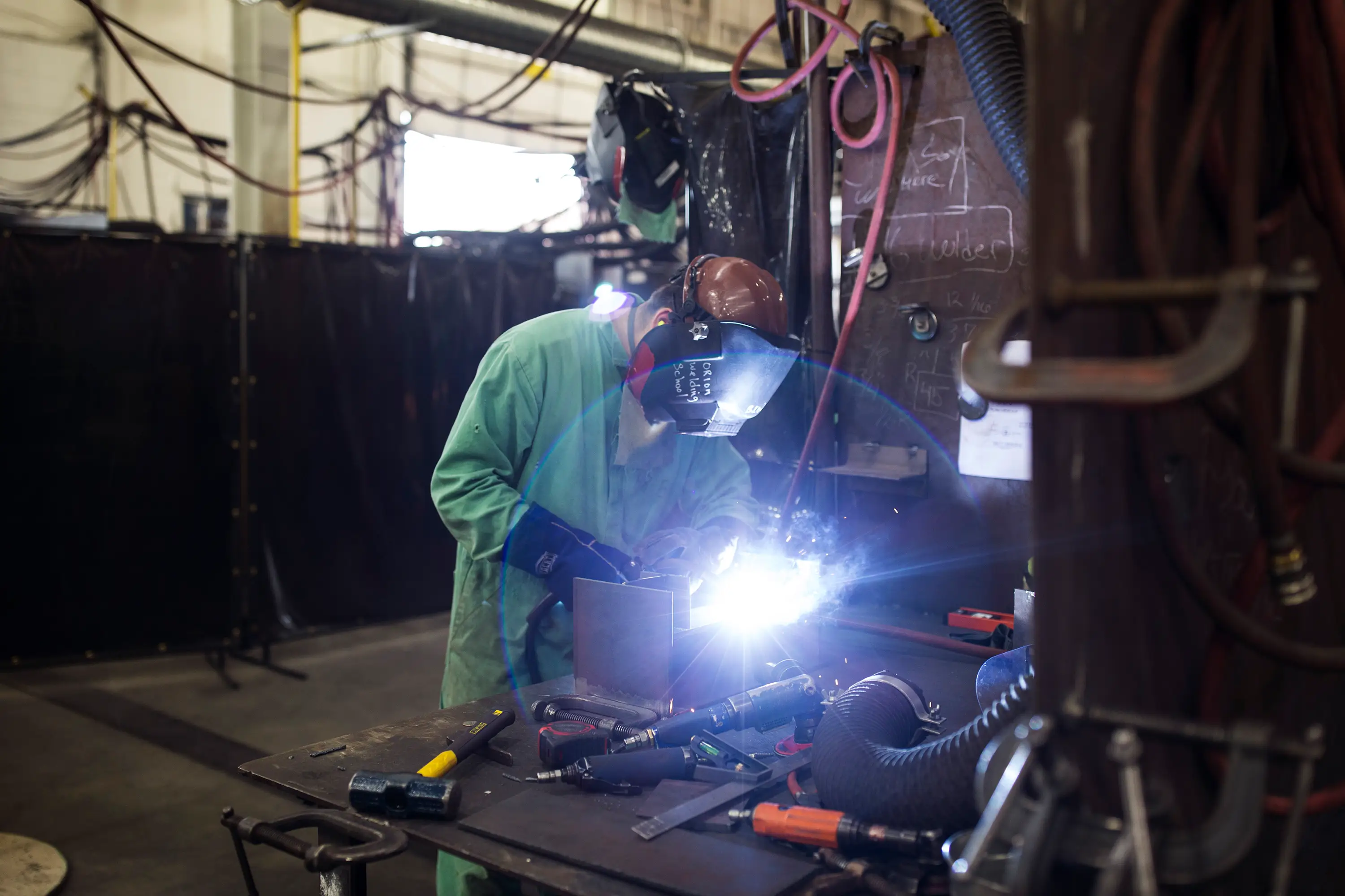 A shipbuilding worker wearing a green worker suit and helmet stands at a work station.