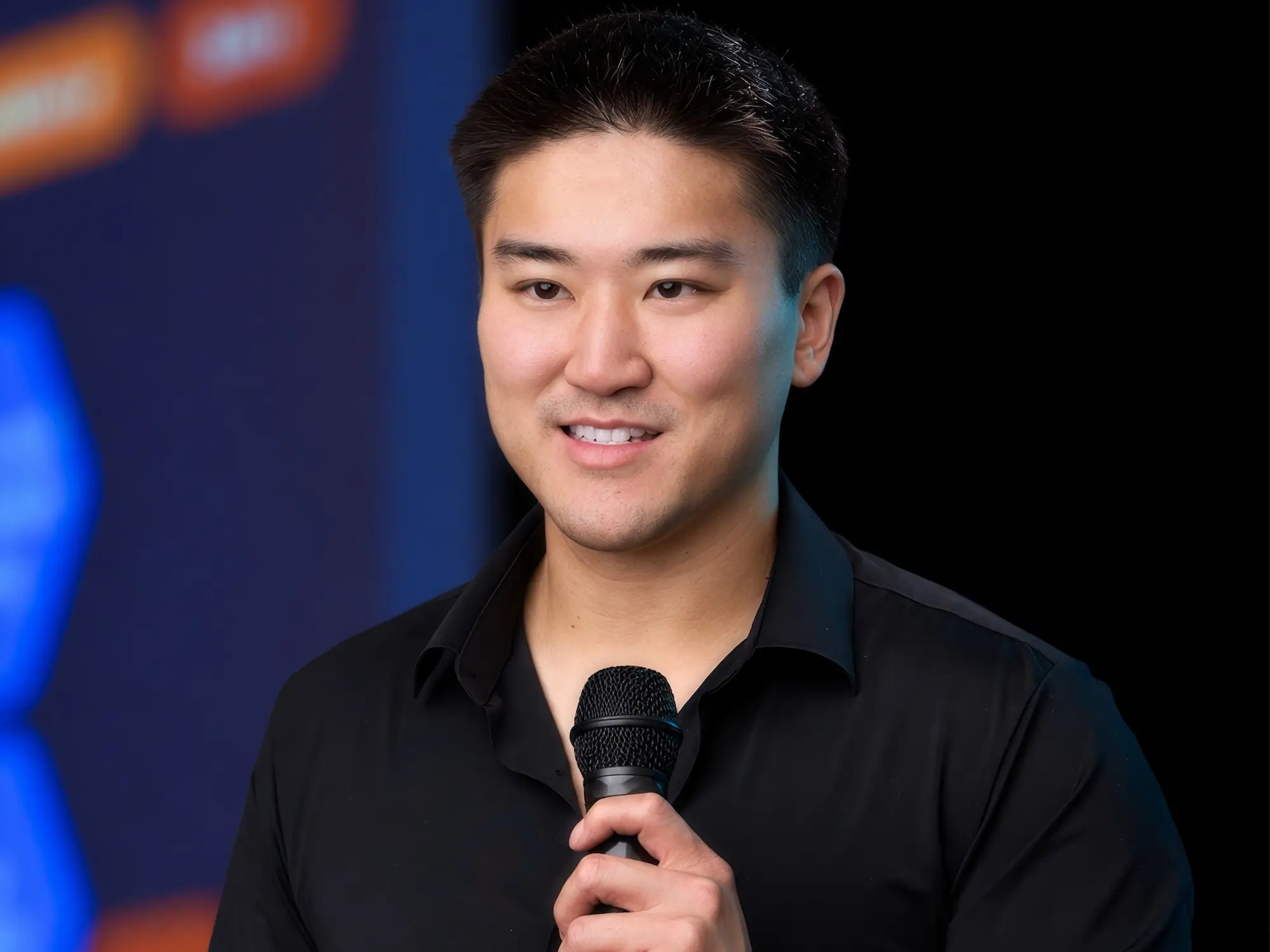 Speaker in a black shirt holds a handheld microphone on a stage with blurred colorful screens.