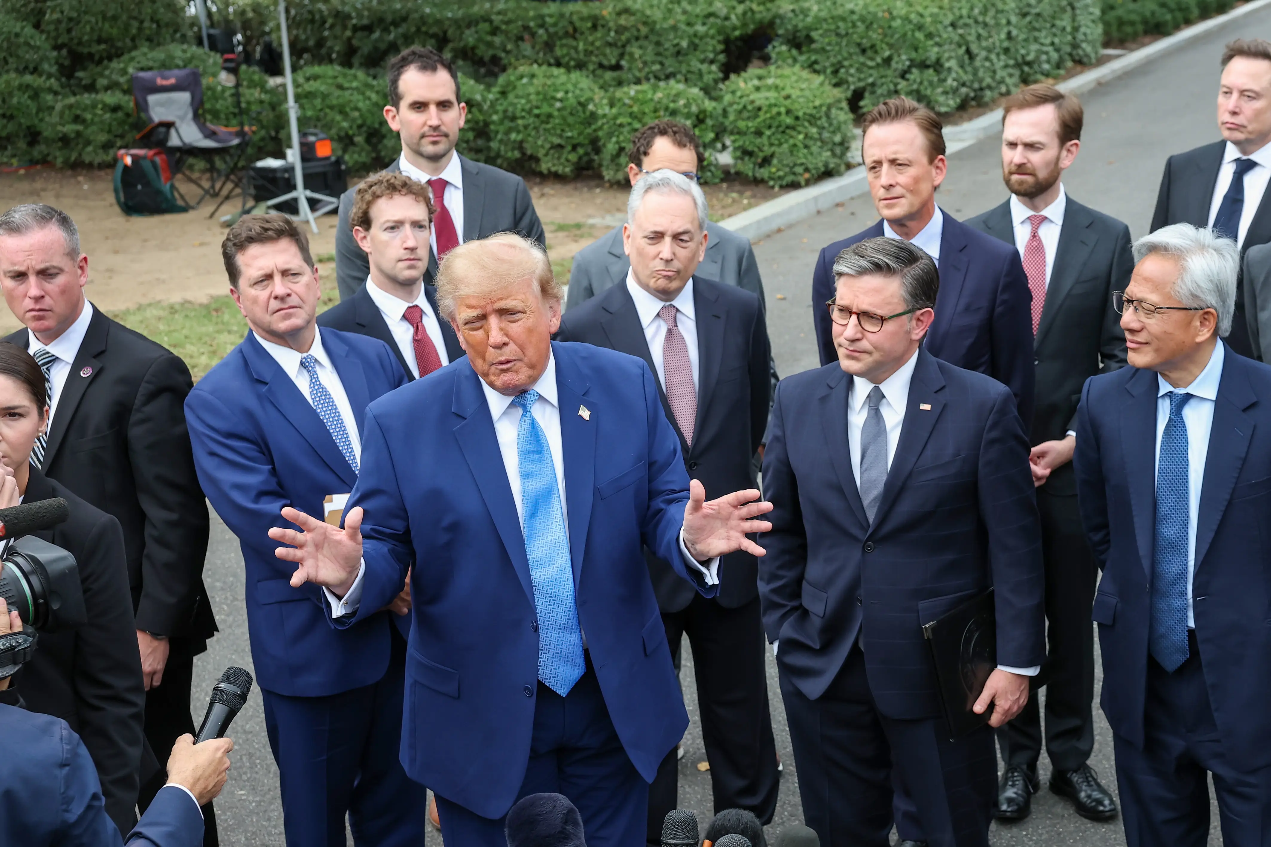 Donald Trump speaks near microphones outdoors with Mike Johnson, Jensen Huang, and other suited attendees nearby.