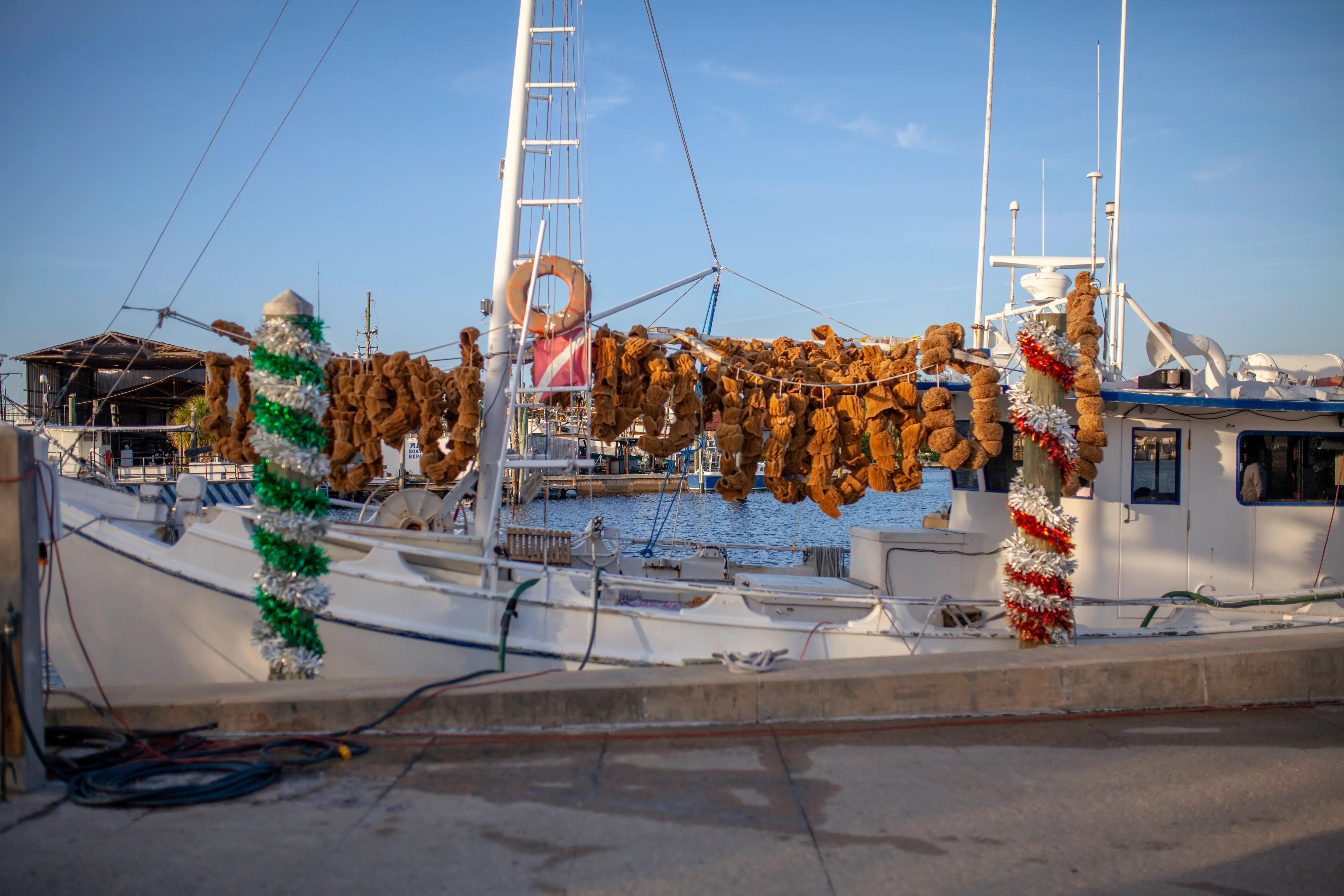 White fishing boat docked in a harbor with sea sponges hanging from rigging and tinsel on poles.