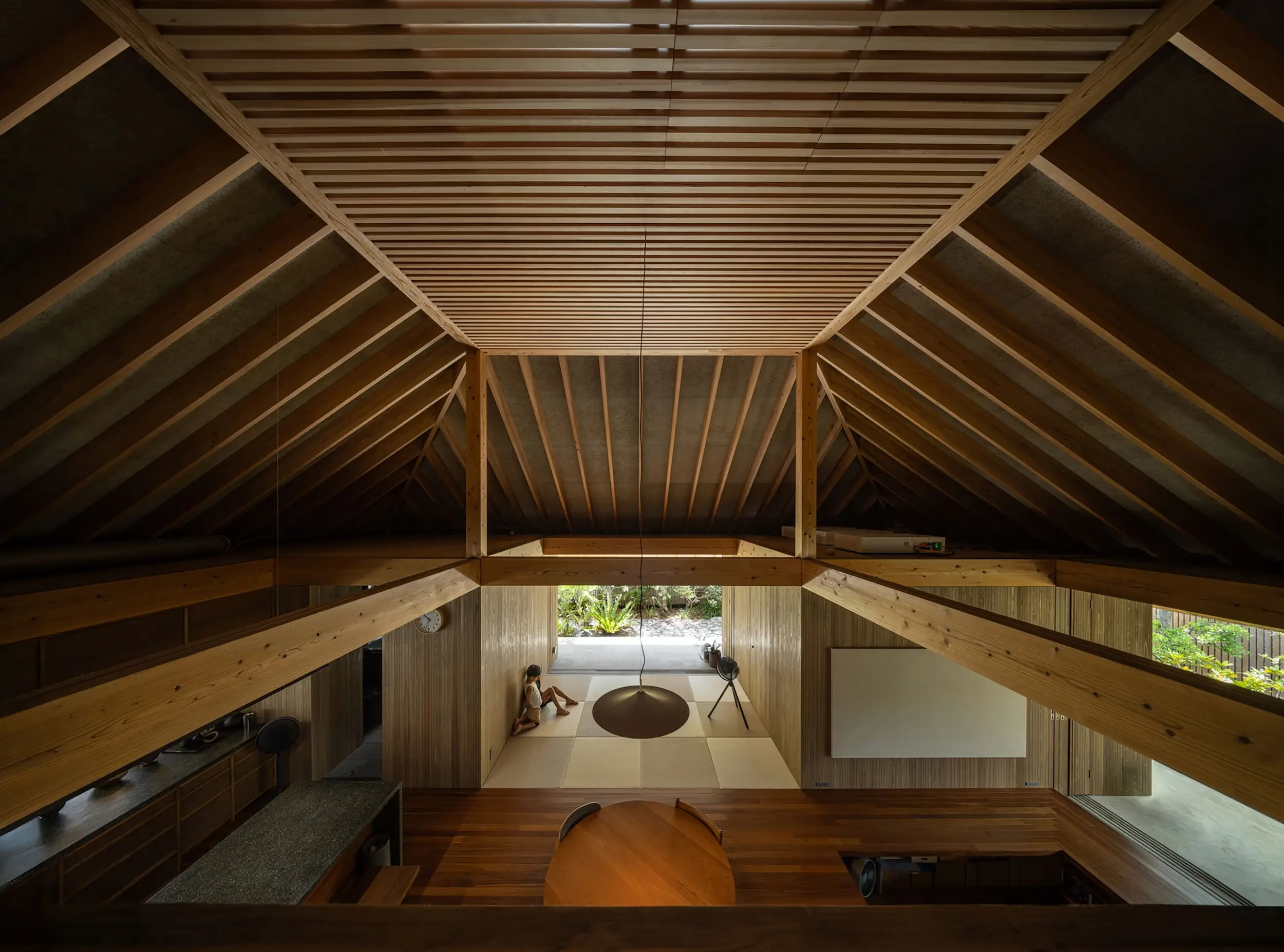 Wooden house interior with exposed rafters, a pendant light, tatami-style sitting area, and garden view.