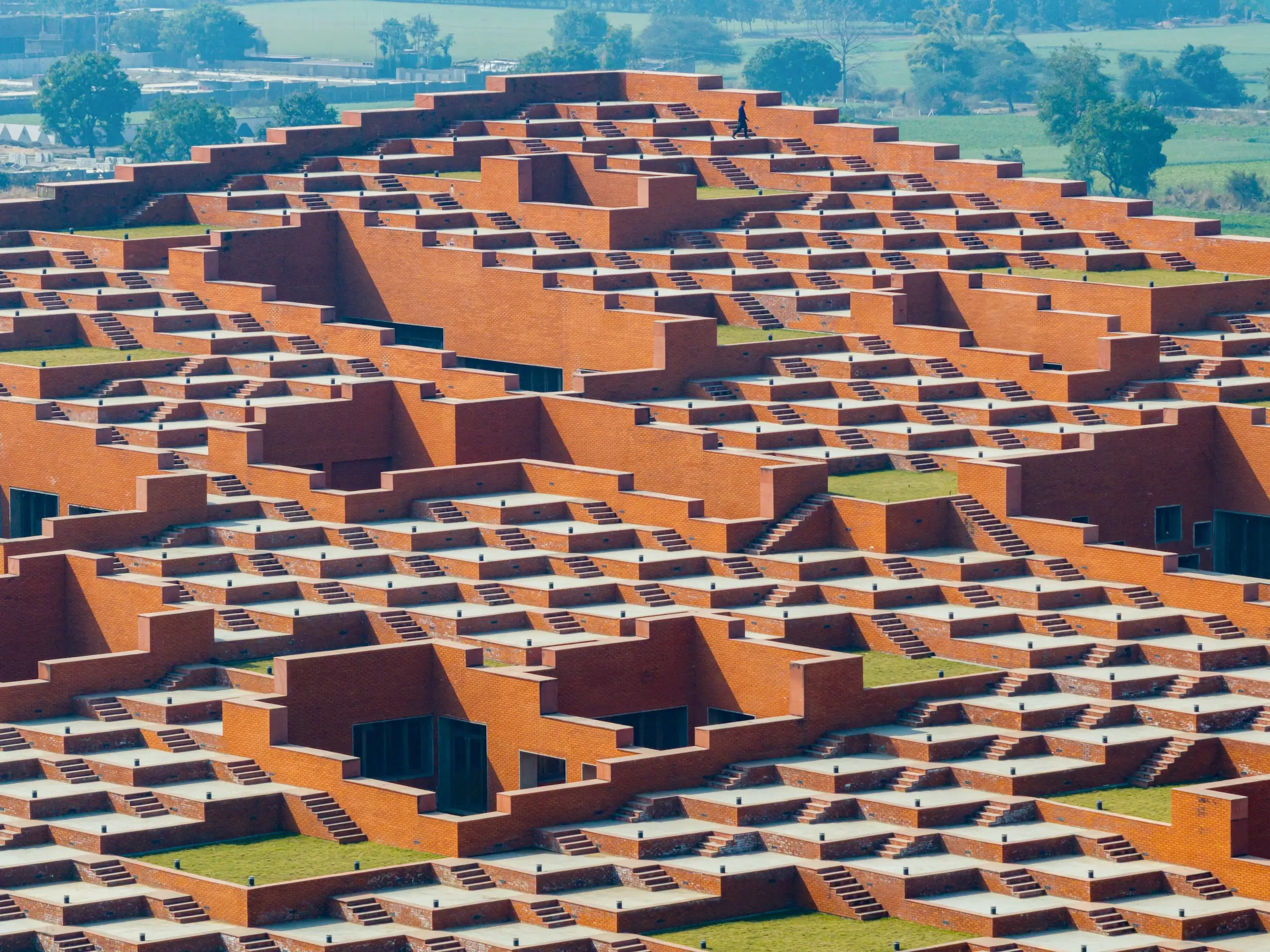 Expansive red brick terraced architectural complex with repeated stairways, courtyards, and lawns amid a green landscape.