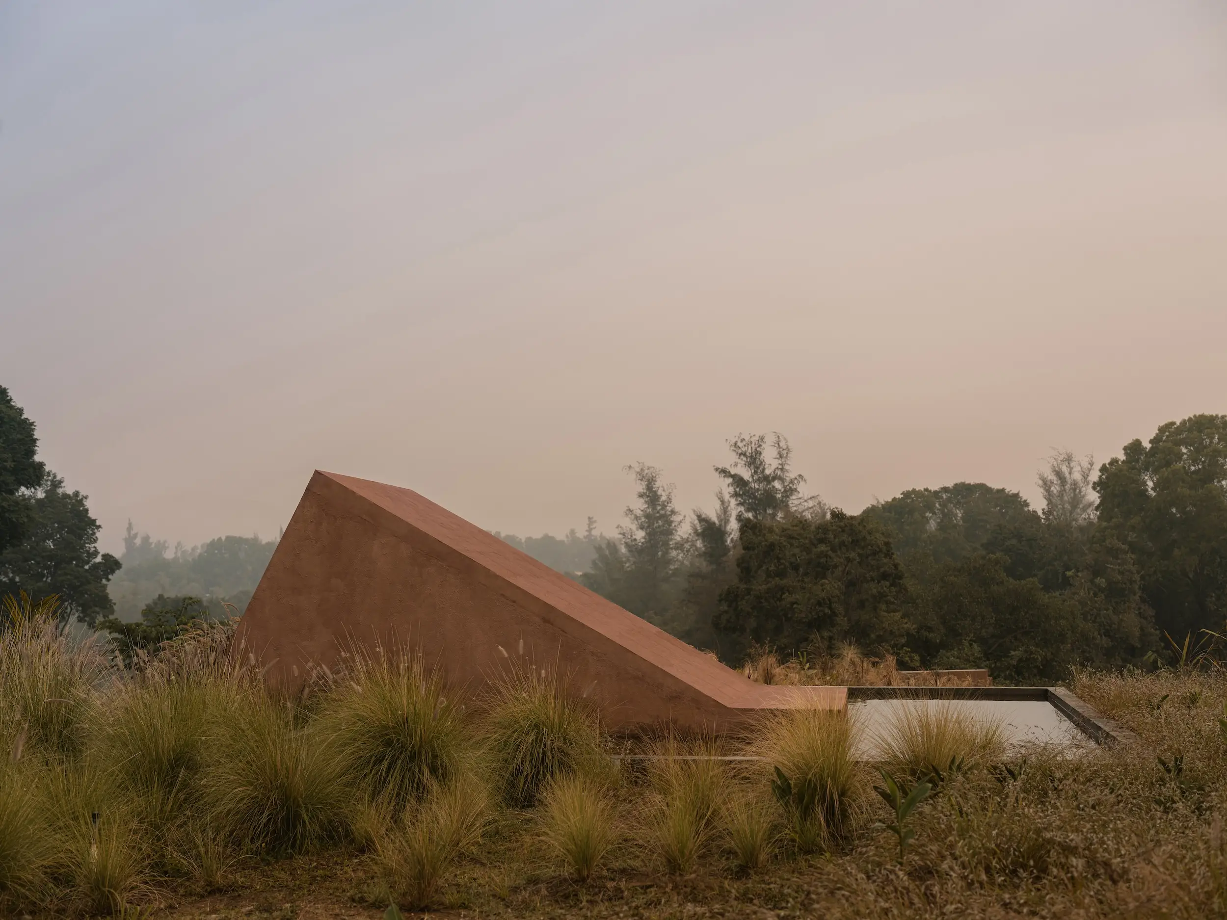 Angular rust-colored concrete structure rises among tall grasses and trees beneath a hazy sky.