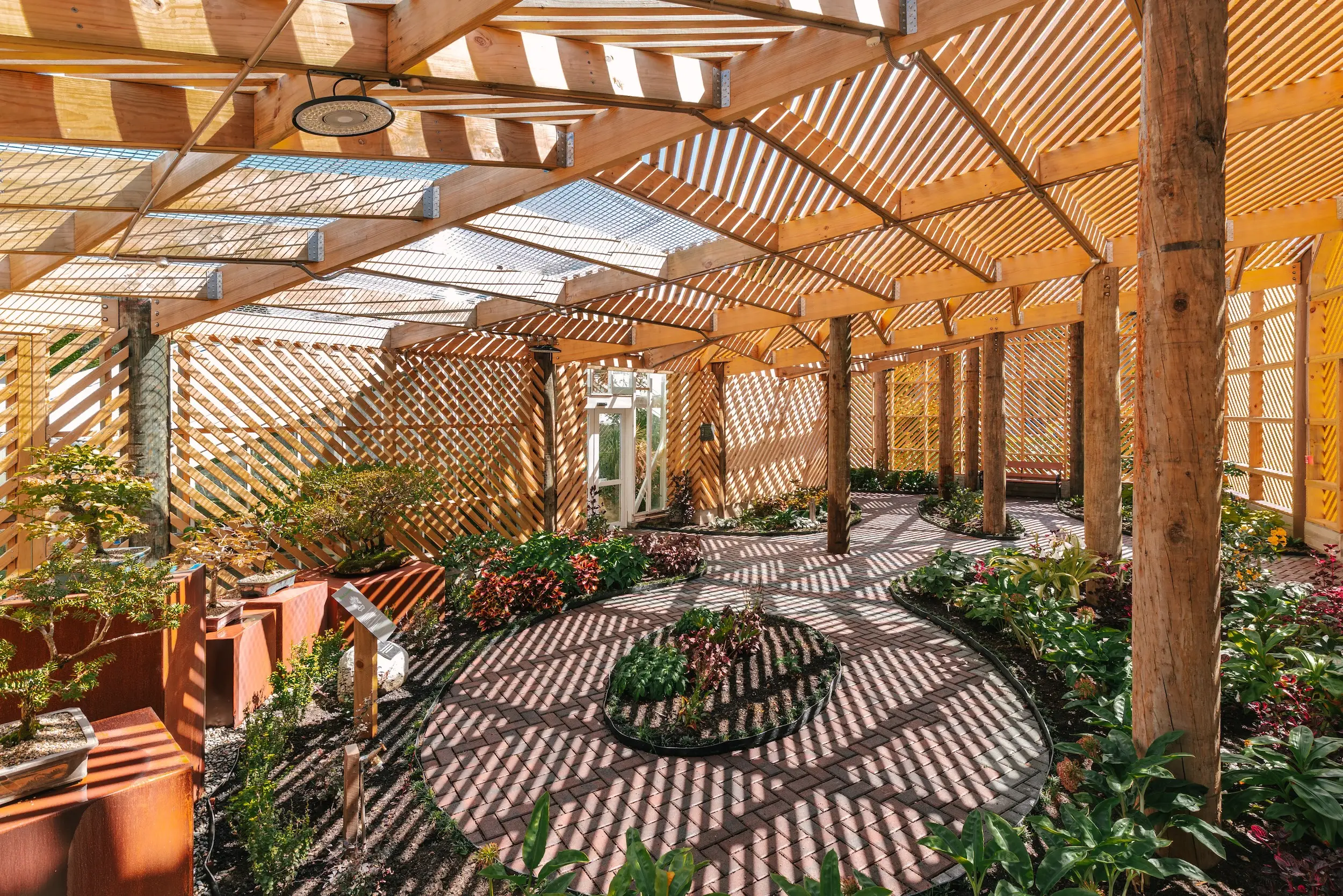 Sunlit indoor garden with wooden lattice walls, beams, brick walkway, planters, and patterned shadows.