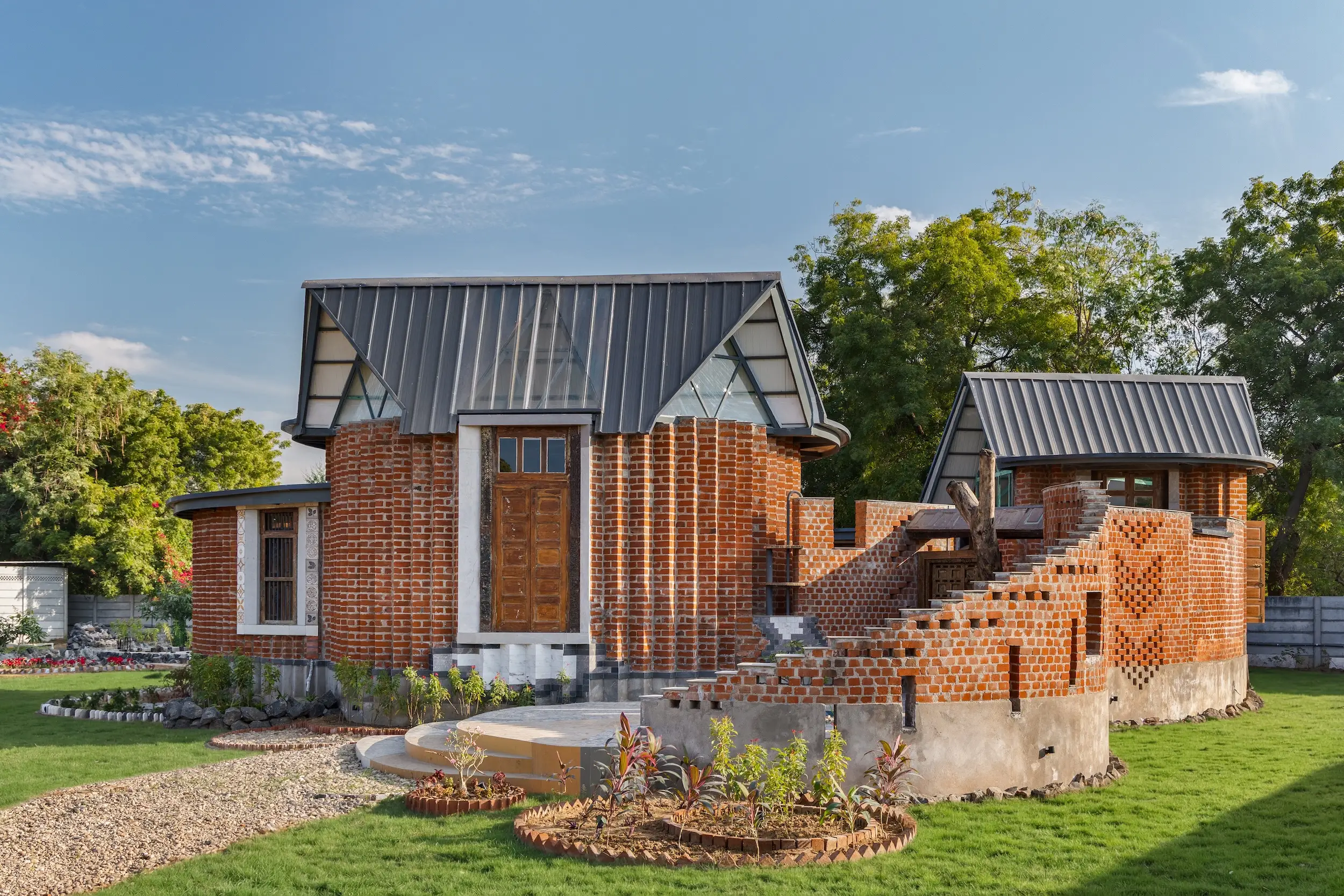 Red brick house with steep gray metal roofs, exterior stairs, landscaped lawn, and trees under a blue sky.