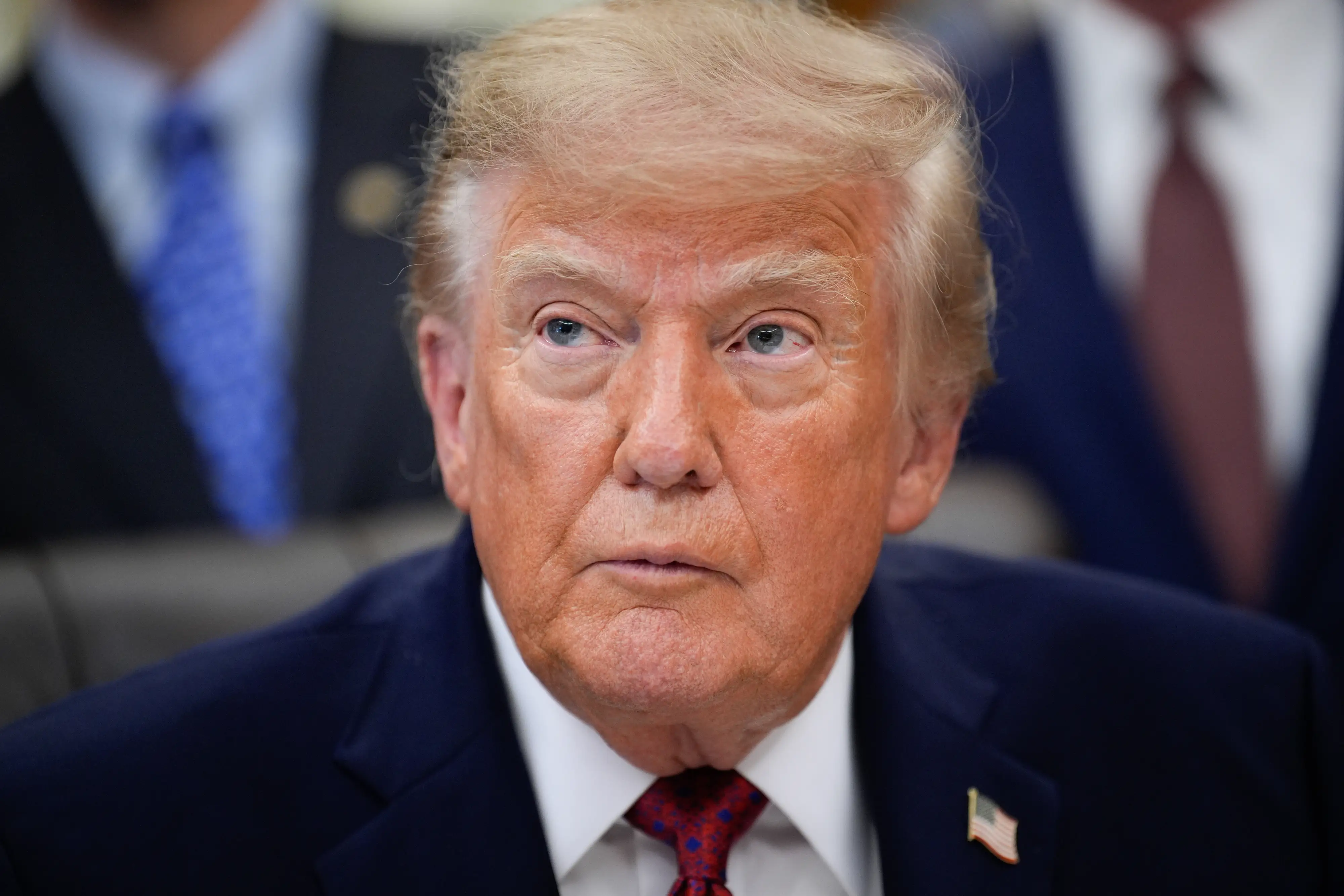 Donald Trump sits at a meeting table wearing a suit, red tie, and American flag lapel pin.