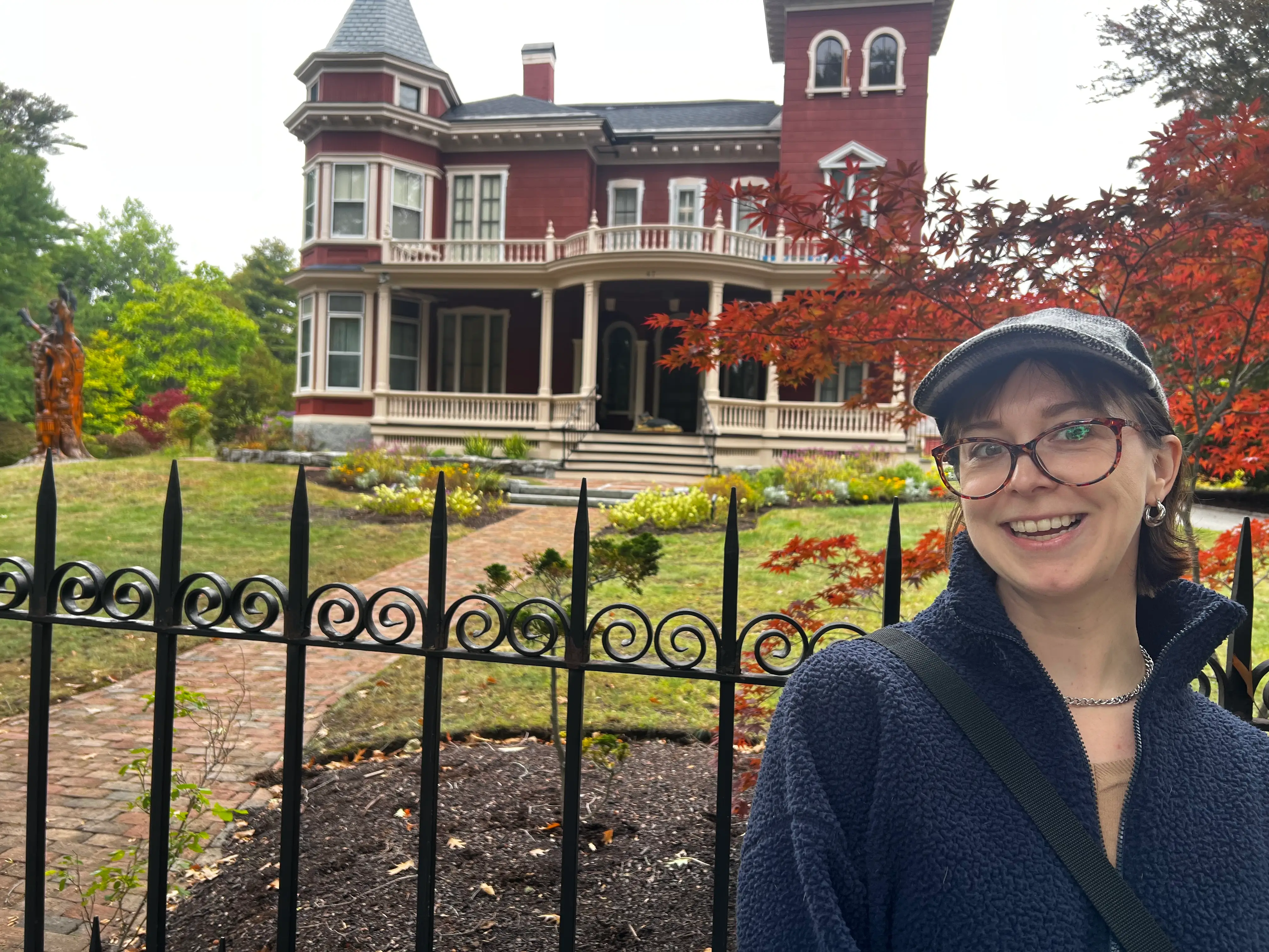 Smiling person in glasses stands by an iron fence before a large red Victorian-style house and autumn trees.