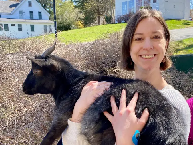 Smiling person in a white sweater holds a black goat on a blue leash outdoors near houses.