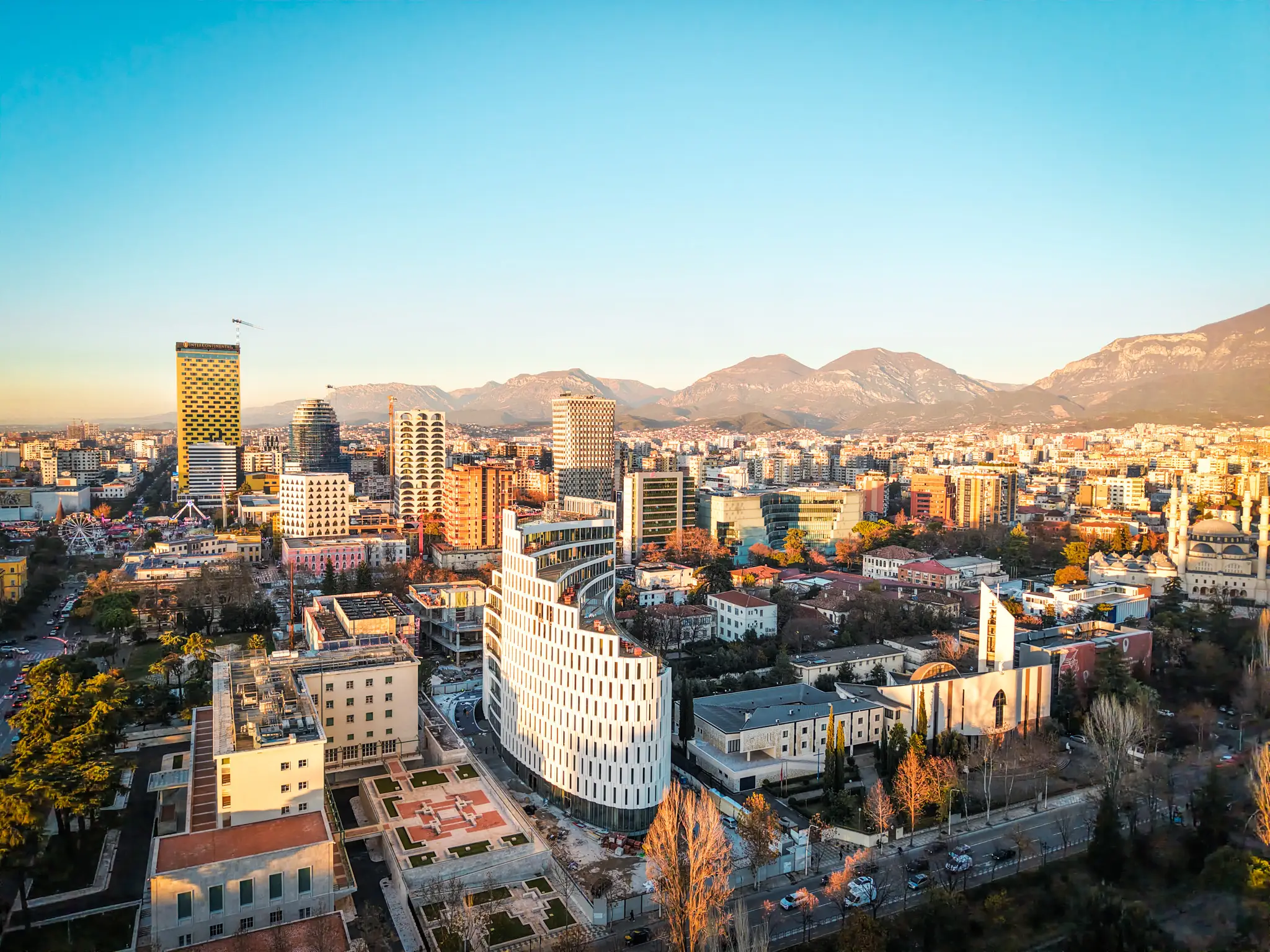 Sunlit Tirana skyline with modern high-rises, a mosque, city streets, and mountains in the background.