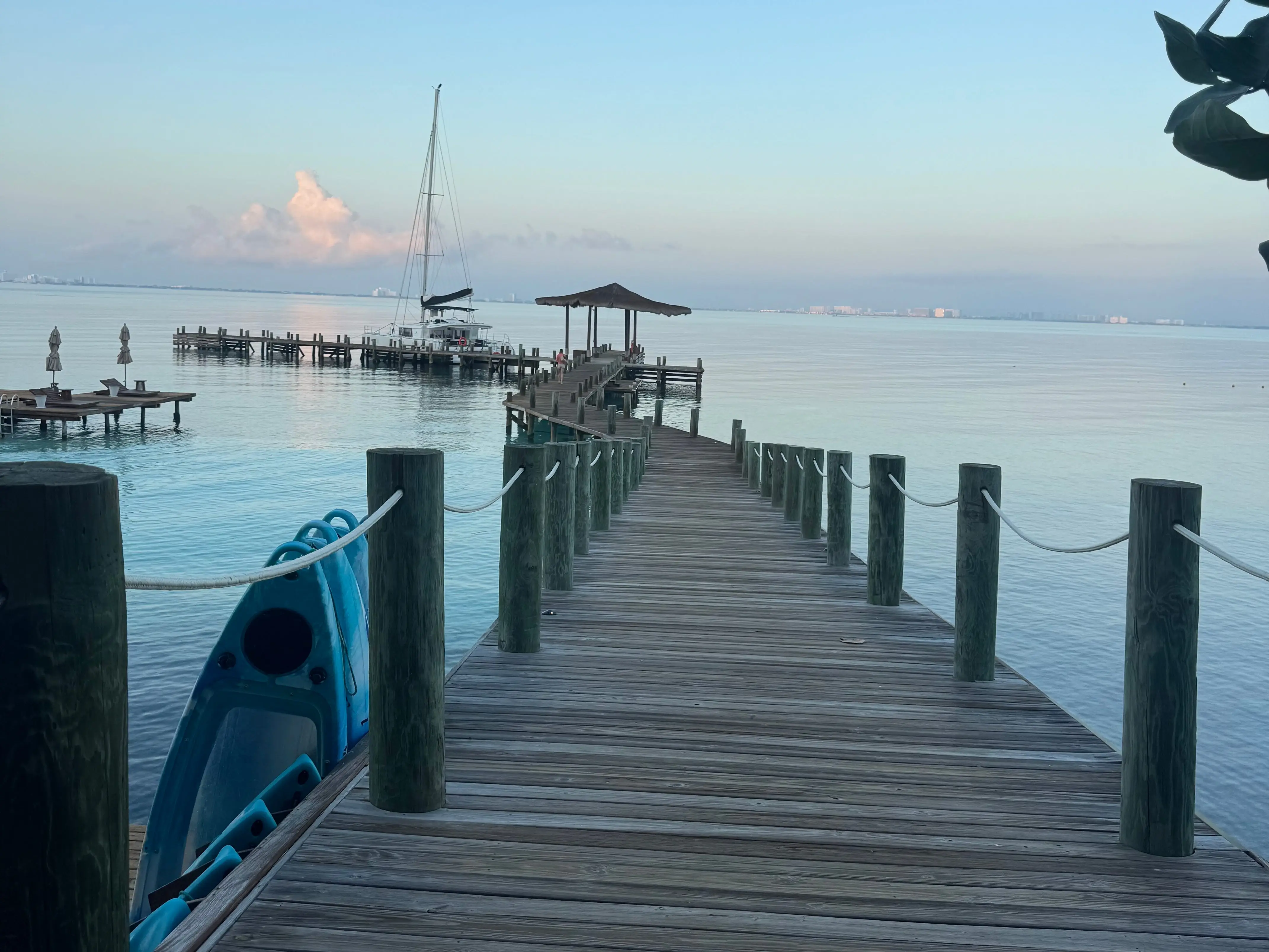 Wooden dock extends over calm coastal water toward a moored sailboat under a clear blue sky on Isla Mujeres.